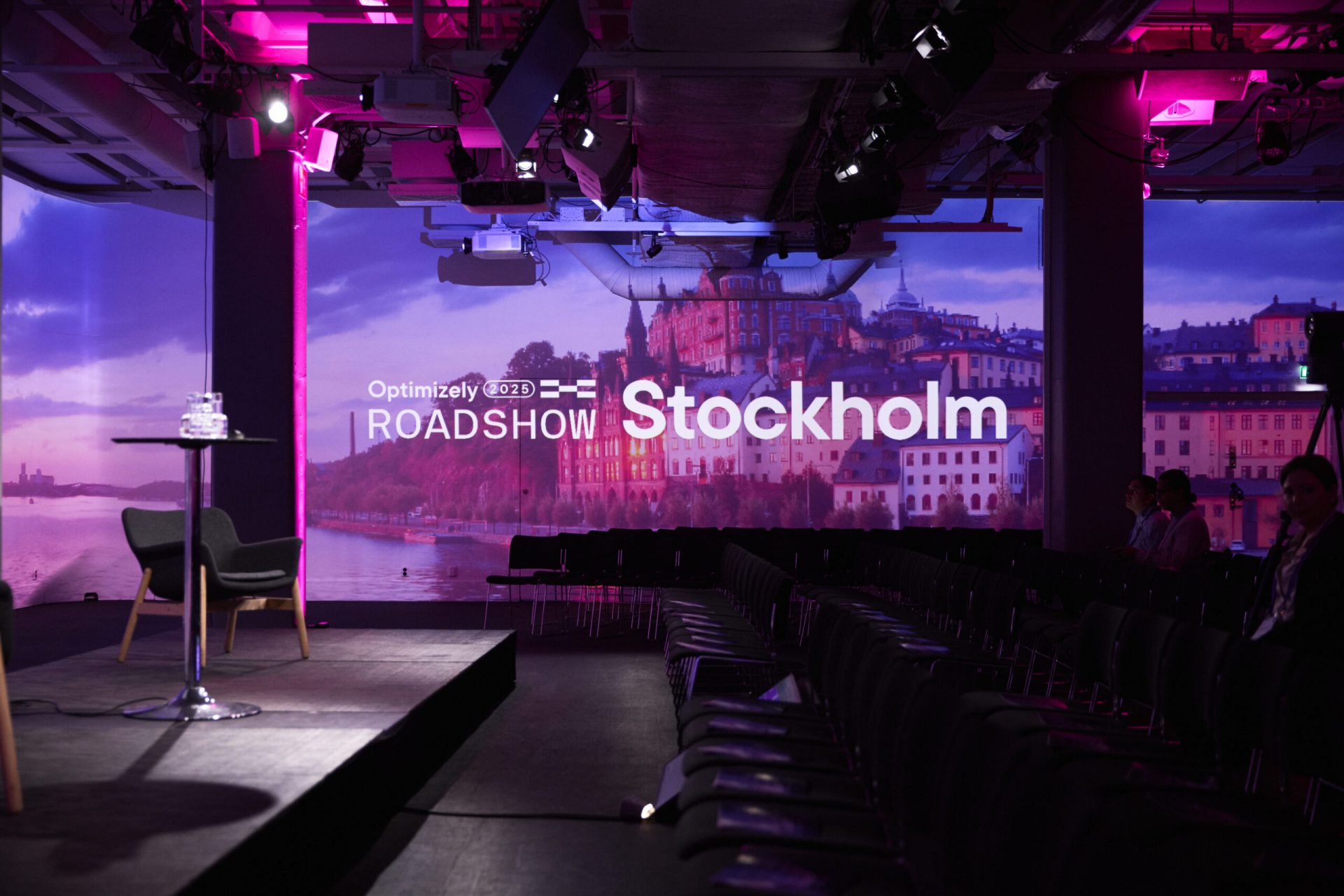 A stage with a chair and small table is set up in front of projected text reading “Optimizely Roadshow Stockholm,” with cityscape images and purple lighting in the background. Rows of empty chairs face the stage.