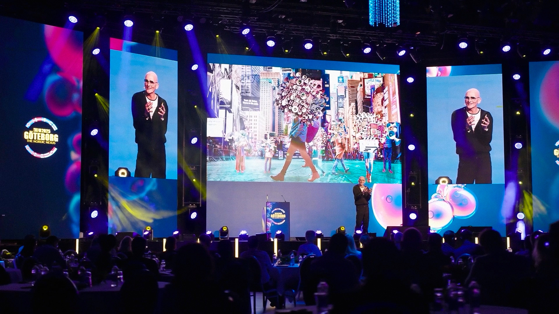 A speaker stands on a stage with large screens displaying his image and colorful graphics, addressing an audience in a dimly lit event venue with blue and purple lighting.