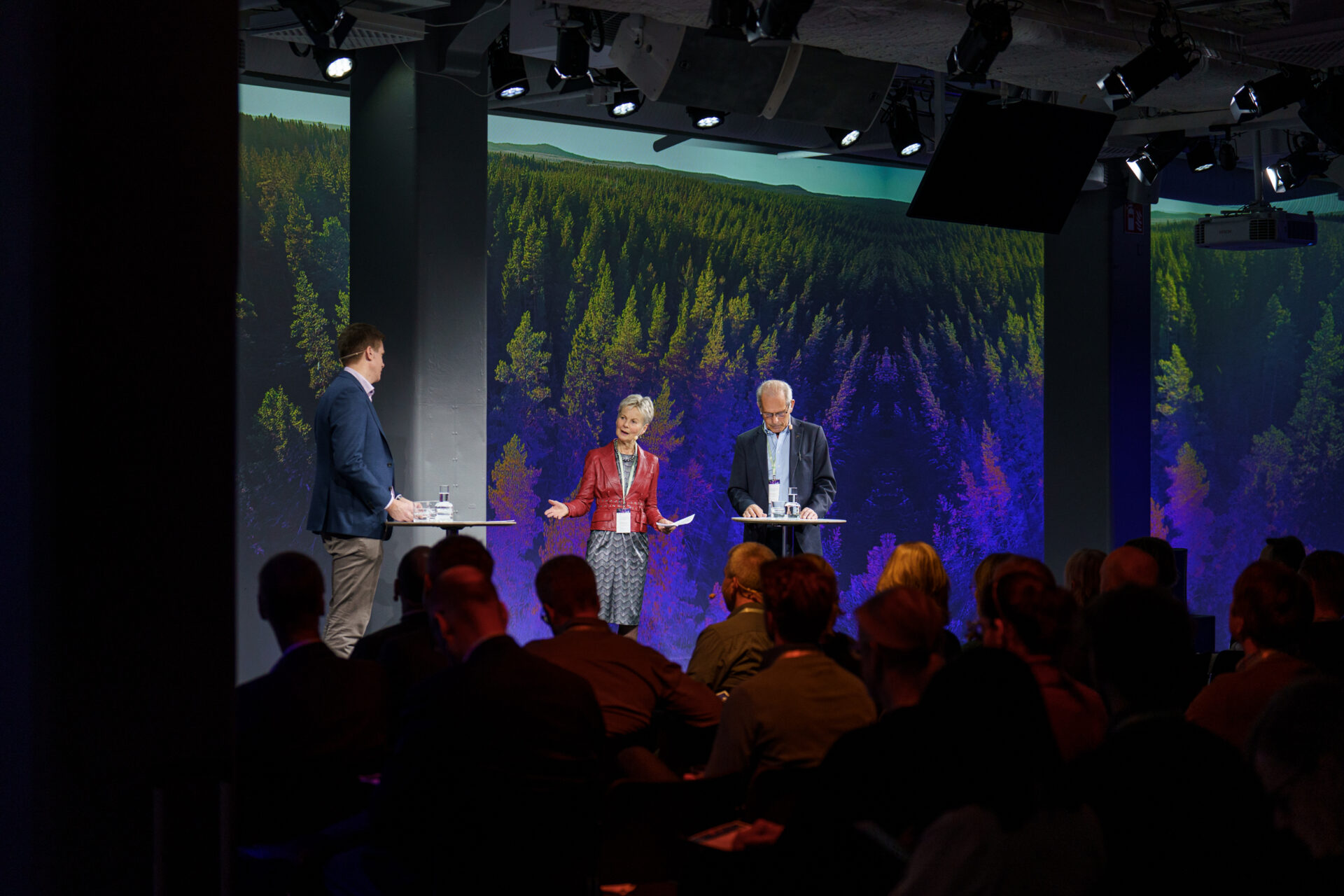 Three people stand and speak on a stage with forest imagery on large screens behind them, addressing an audience seated in front of them in a darkened room.