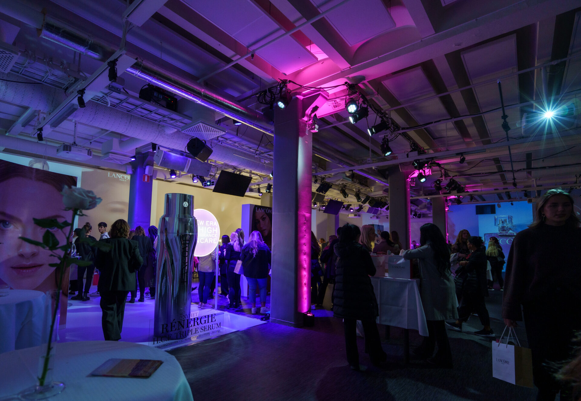 A modern event space with purple and pink lighting, people mingling near displays and tables, and large promotional posters on the walls. A single white rose in a vase sits on a table in the foreground.