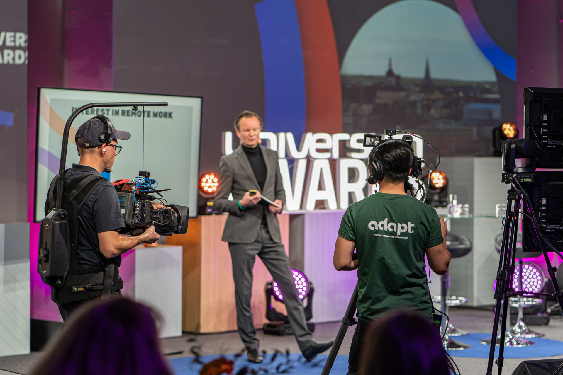 A man in a suit stands on a stage with a microphone, facing cameras and crew members recording him. Behind him is a screen that says “UNIVERS” and studio lights, in a professional studio setting.