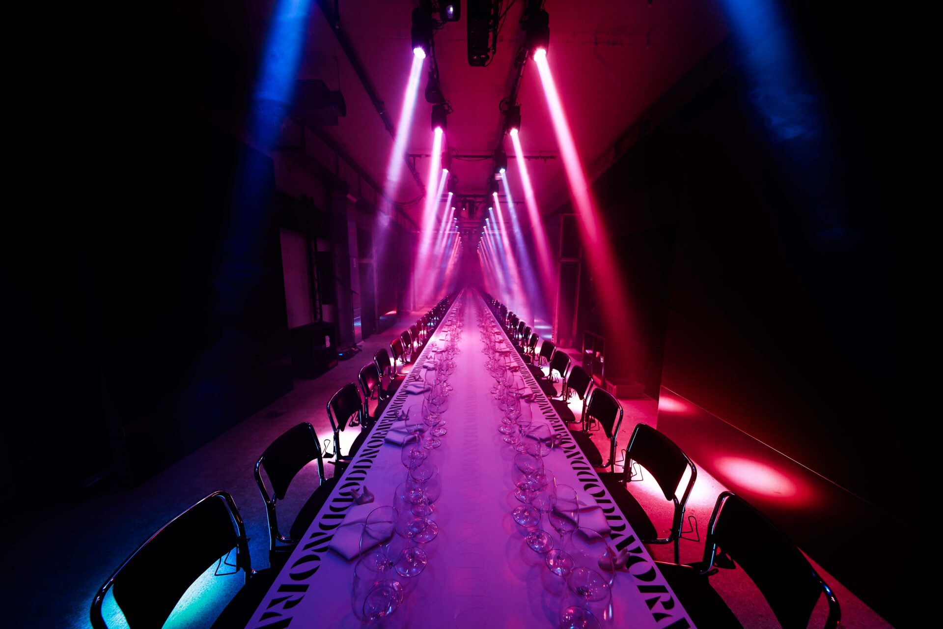 A long, elegant dining table set with glassware and napkins is illuminated by dramatic pink and blue stage lights, creating a vibrant, futuristic atmosphere in a dark room.