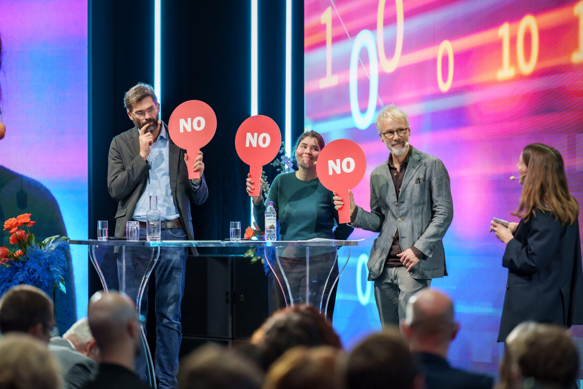 Three people stand behind a clear table on stage, each holding a red paddle with NO written on it. They face a woman who stands to the right, while an audience watches. The background is lit with colorful digital graphics.