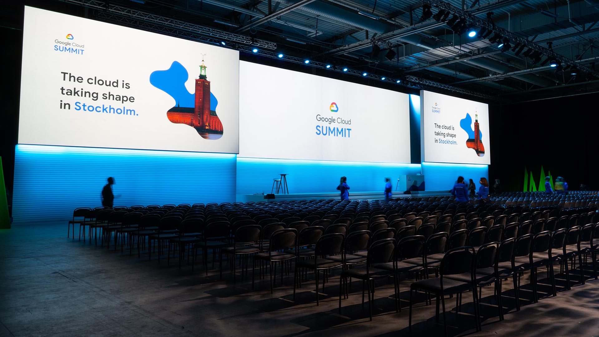 Rows of empty chairs face large screens at a Google Cloud Summit event. The screen reads “The cloud is taking shape in Stockholm.” A few people walk around the venue under blue lighting.
