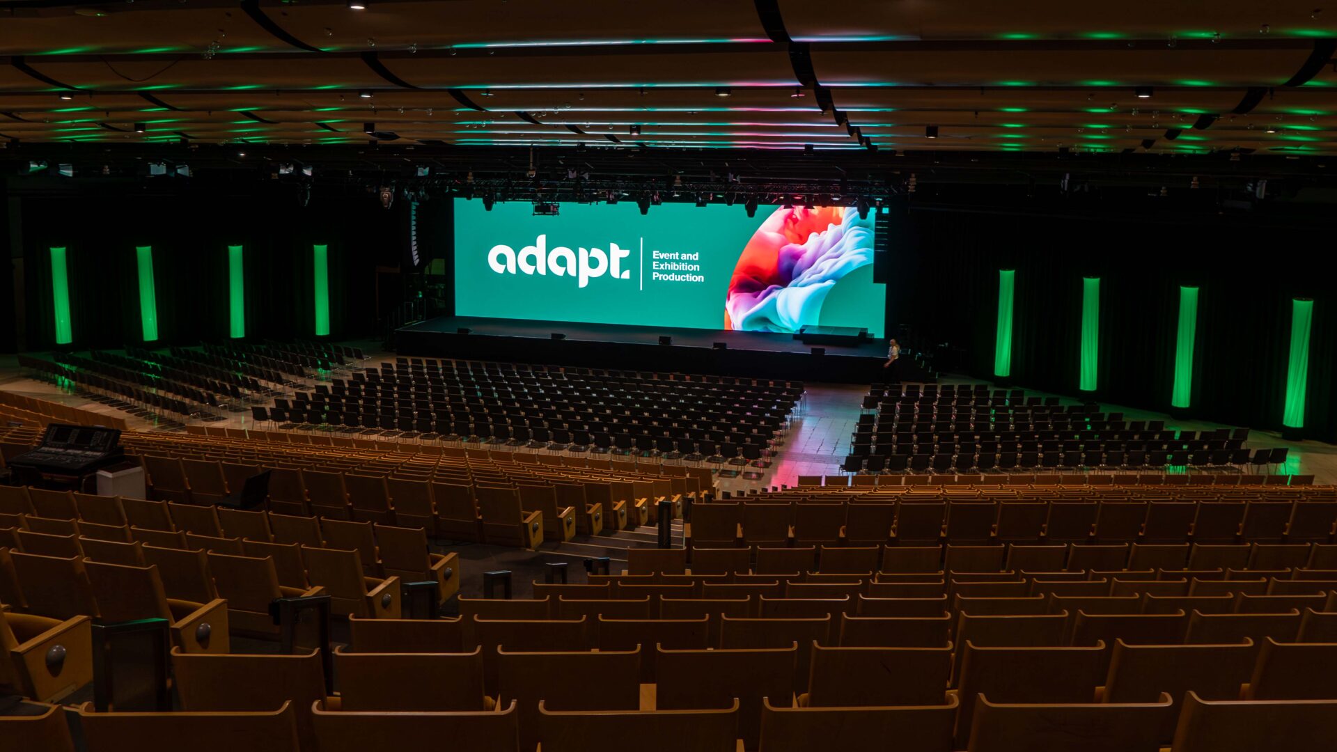 A large, empty auditorium with rows of wooden seats facing a brightly lit stage displaying “adapt” and colorful graphics on screens. Green vertical lights illuminate the walls and ceiling.
