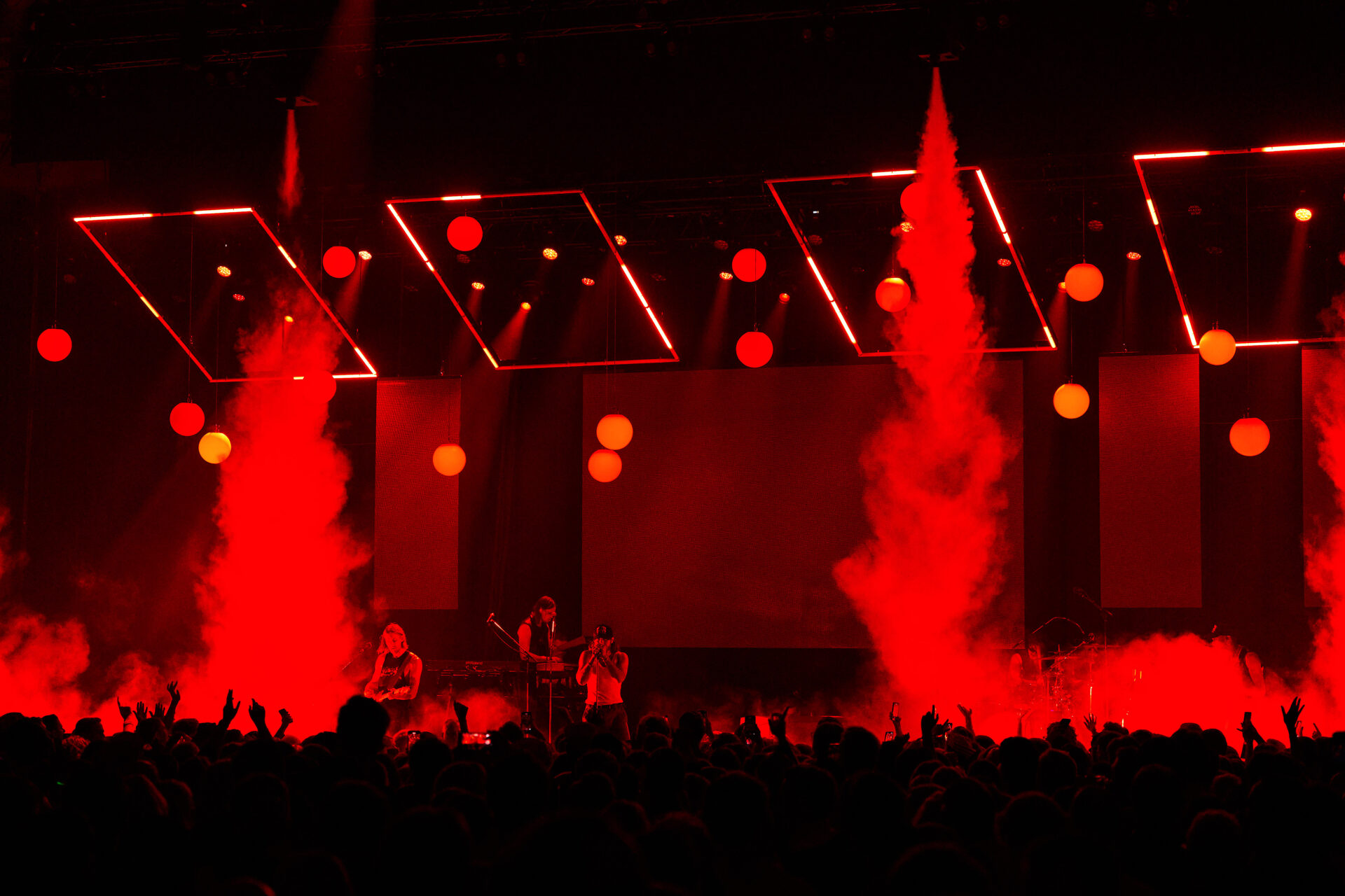A concert stage lit with red lights features performers and dramatic red smoke effects, with geometric light structures above and a crowd of people silhouetted in the foreground.