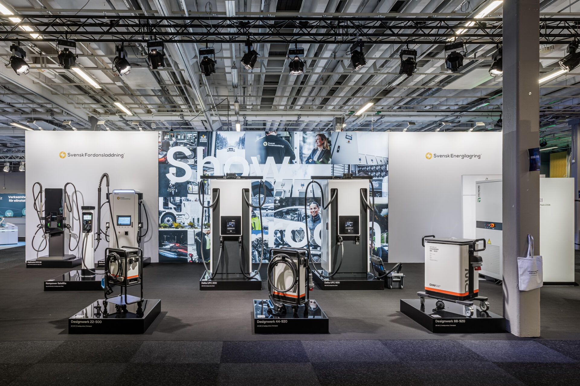 A trade show booth displays multiple electric vehicle chargers and charging equipment on black platforms with cables, set against a backdrop featuring technology images and the words Show. Overhead lights illuminate the scene.