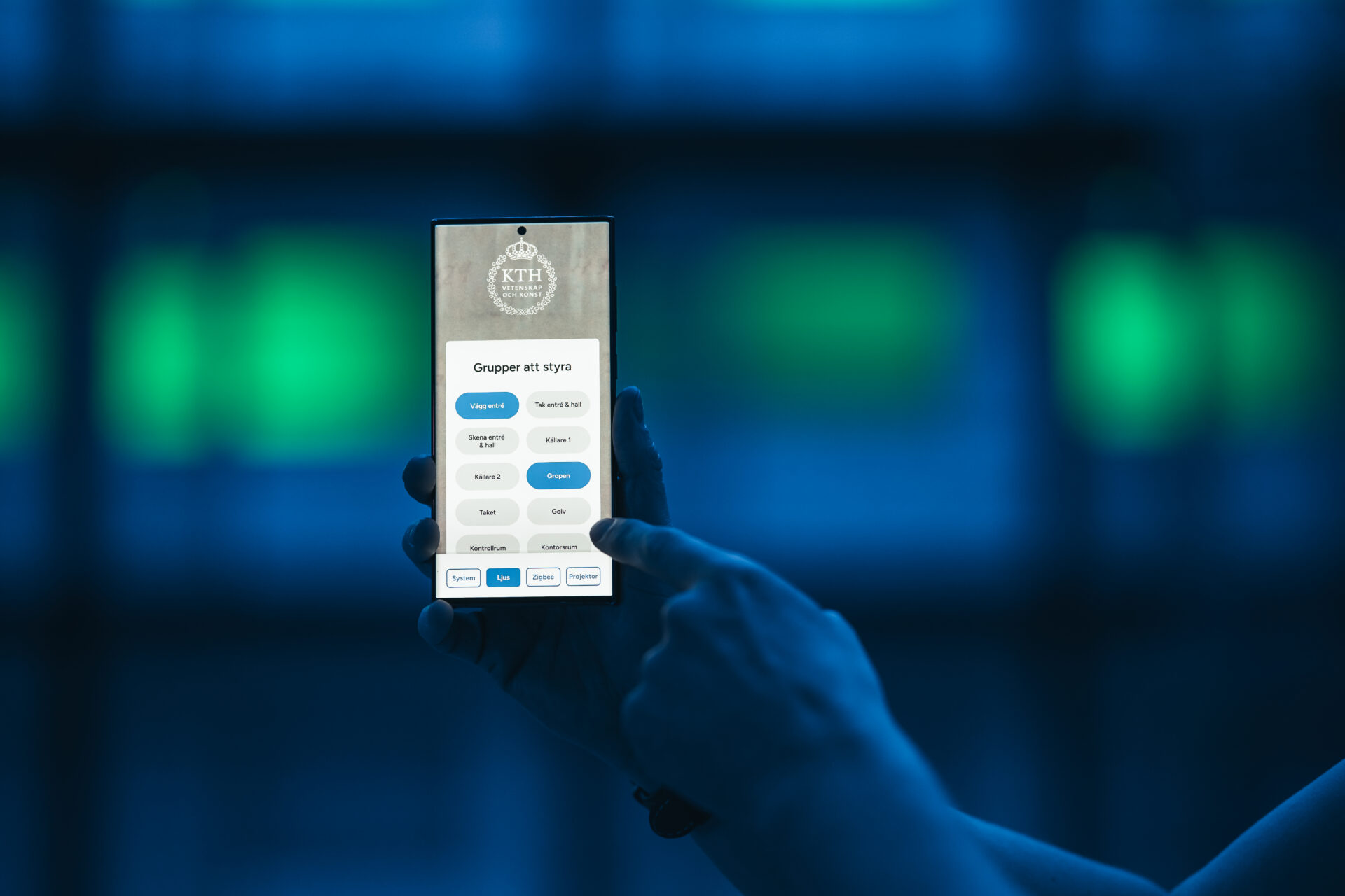A person holds a smartphone with a Swedish app on the screen. The app displays the KTH Royal Institute of Technology logo and several selectable groups or categories. The background is blurred and blue-toned.