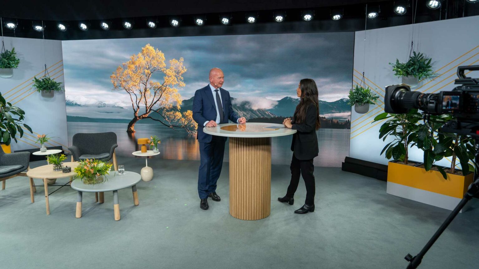 A man in a suit and a woman in black attire stand at a round table on a modern studio set, with chairs, plants, and a large scenic backdrop of a tree by a lake and mountains behind them. A camera is visible on the right.
