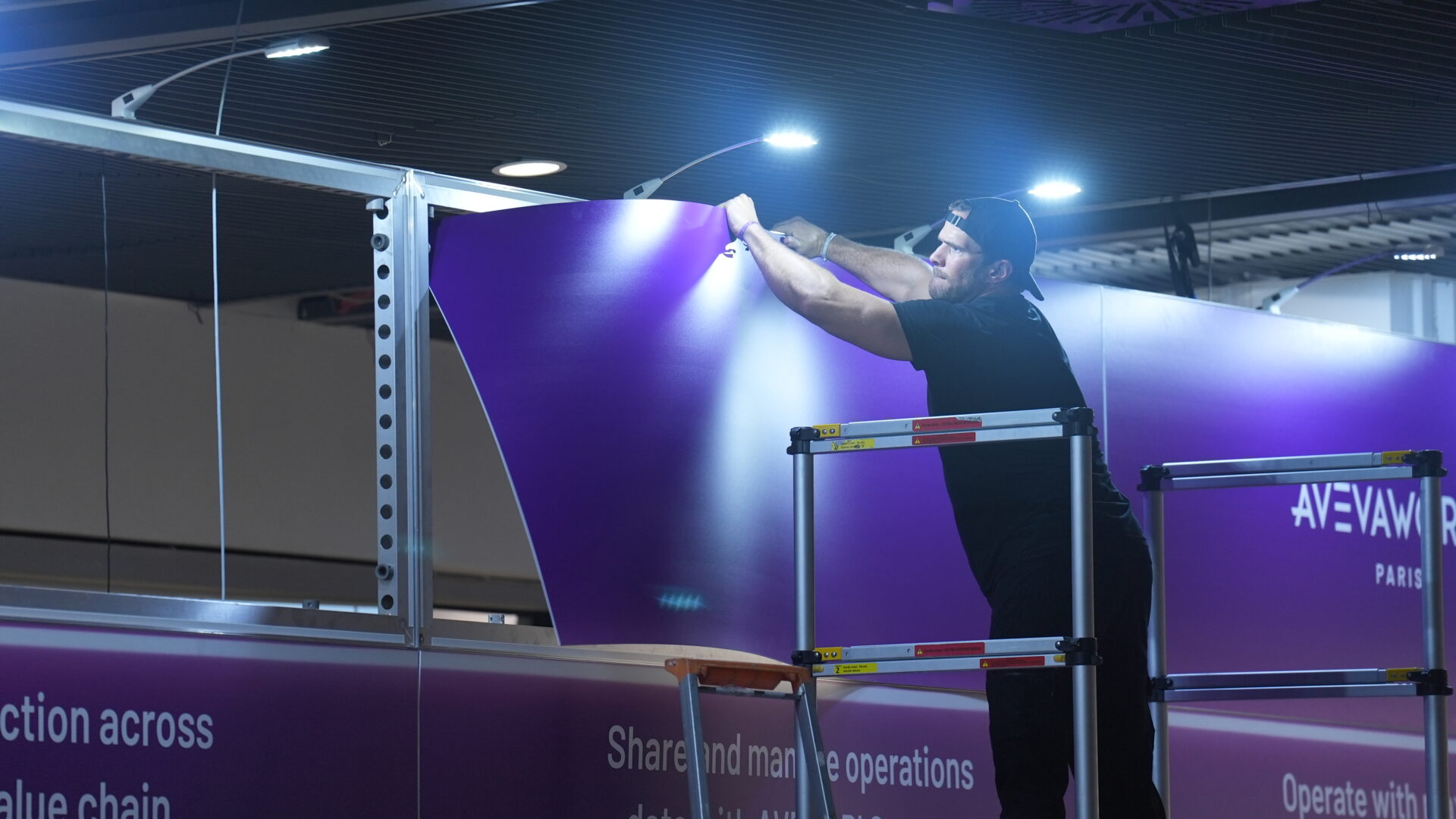 A man standing on a platform installs a large purple sign at an indoor event or trade show, using both hands to attach it to a metal frame under bright overhead lights.