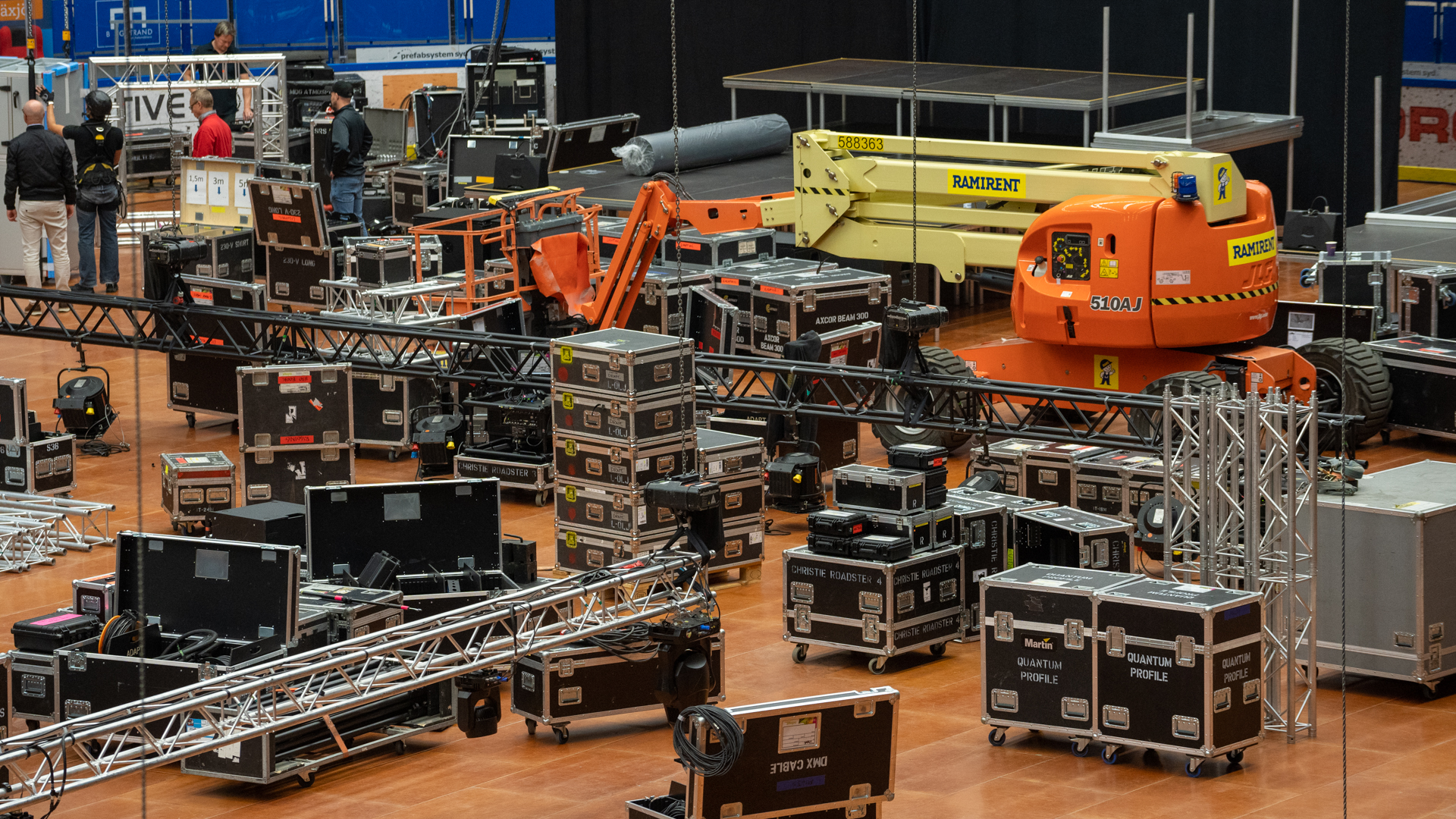 Stage equipment and lighting cases, metal trusses, and an orange scissor lift are spread across an indoor venue floor. Several workers are setting up for an event or concert in the background.