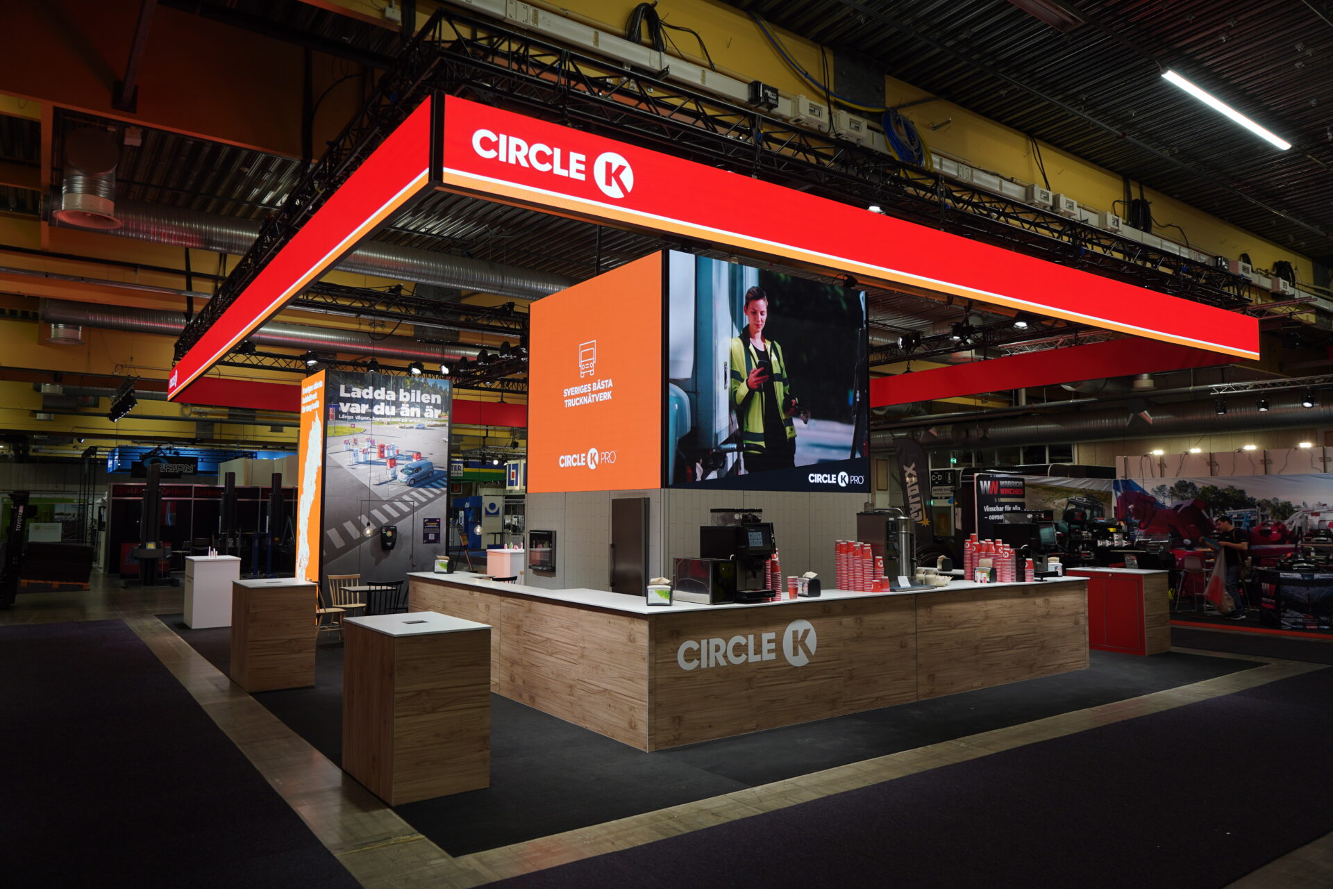 A modern Circle K exhibition booth with wooden counters, digital screens displaying promotional content, and branded signage, set up in a large, well-lit indoor space.