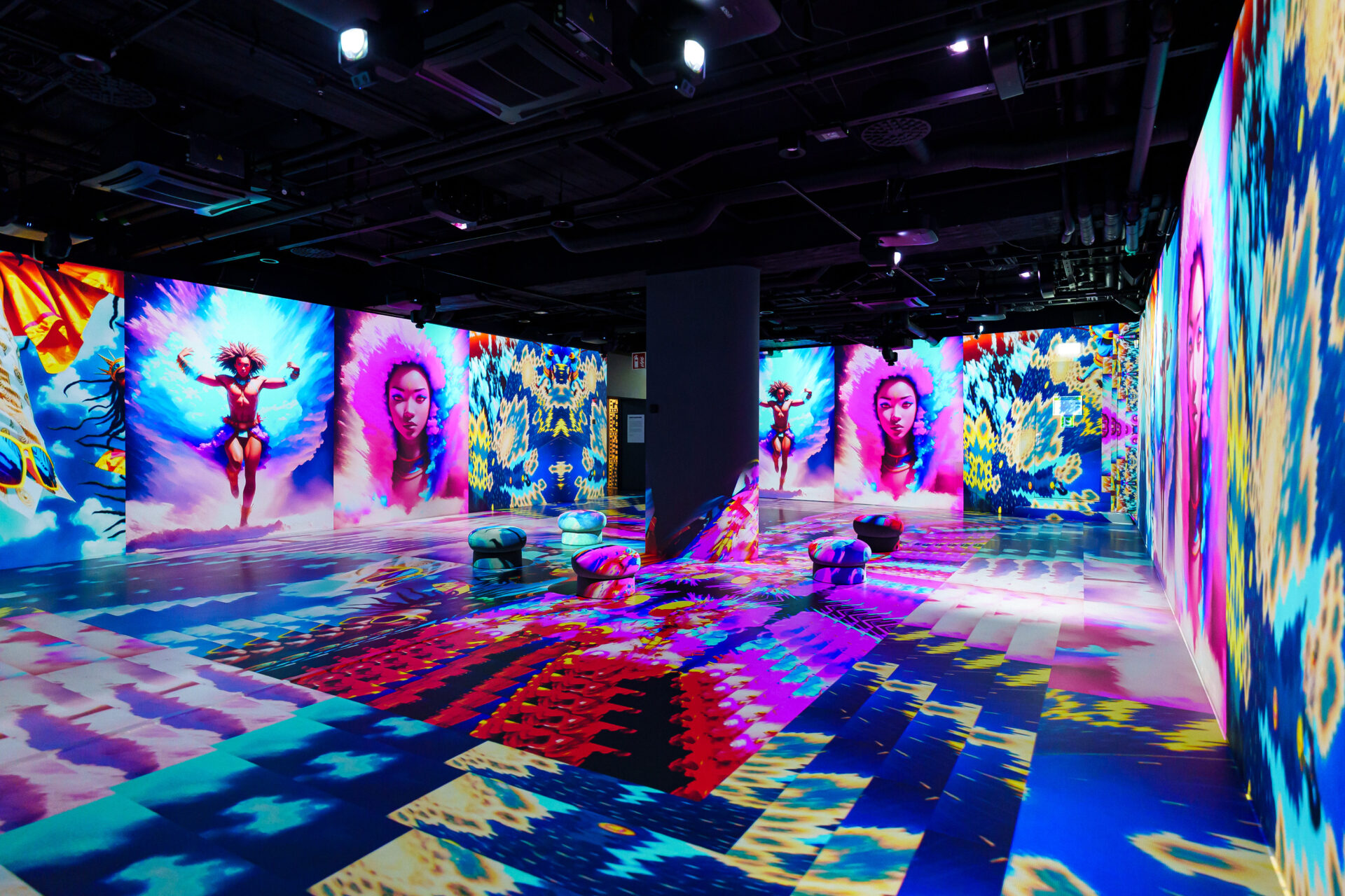 A colorful immersive art installation features vivid projections of a woman with feathered wings on the walls, mirrored patterns on the floor, and several cushioned seats scattered throughout the vibrant room.