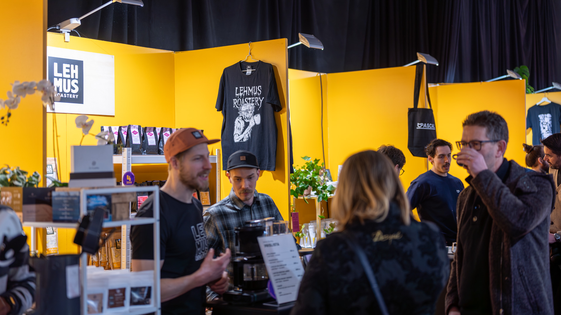 Several people interact and browse products at a brightly lit booth with yellow walls labeled Lehmus Roastery. Coffee-related items and shirts are displayed, and a barista serves customers.