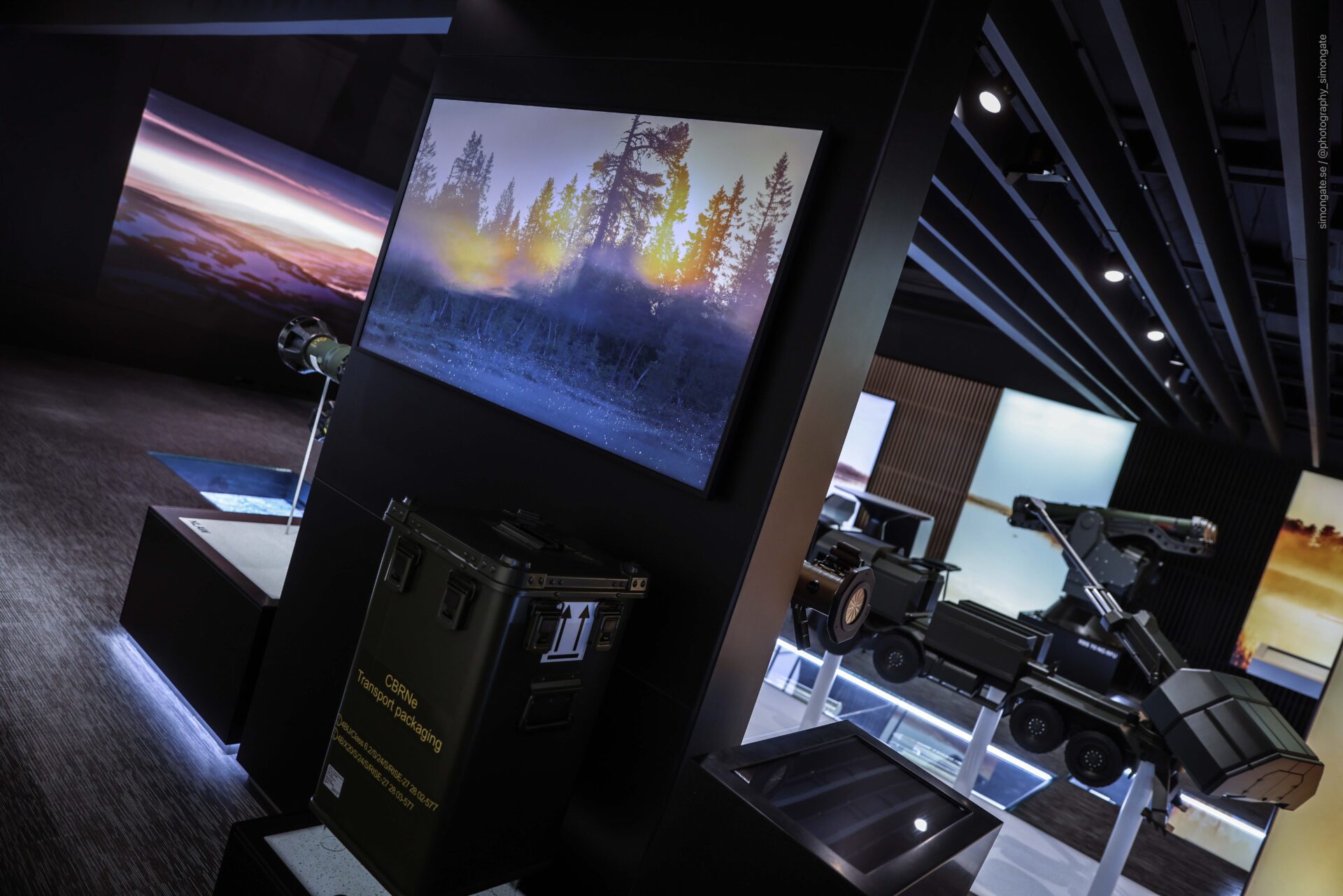 A modern exhibition space displays military equipment, including remote-controlled vehicles and cases, with illuminated screens showing nature and landscape images on dark walls.