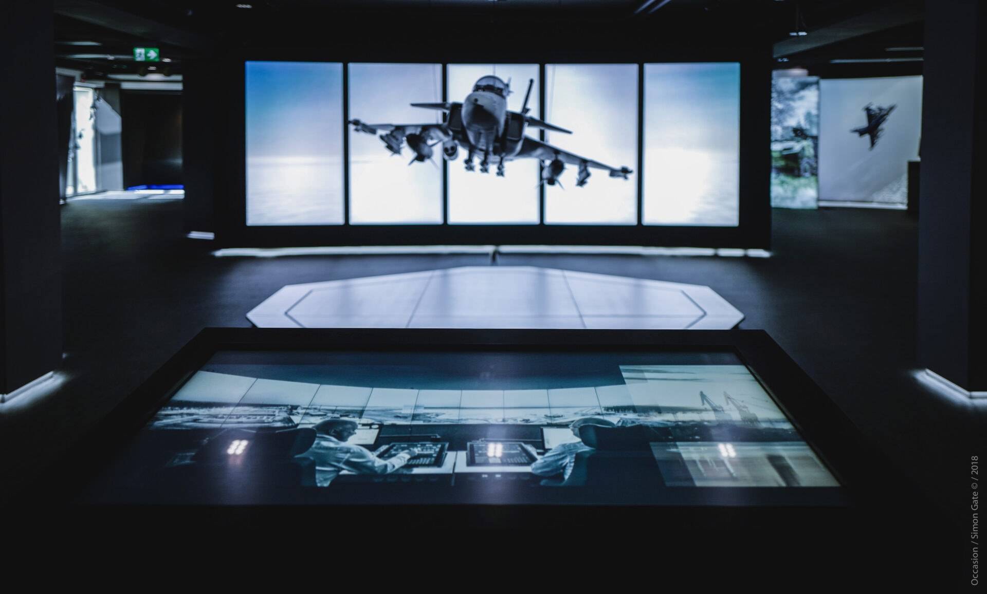 A dark exhibition space features a large screen showing a fighter jet in flight. In the foreground, another screen displays two people operating equipment in a control room.
