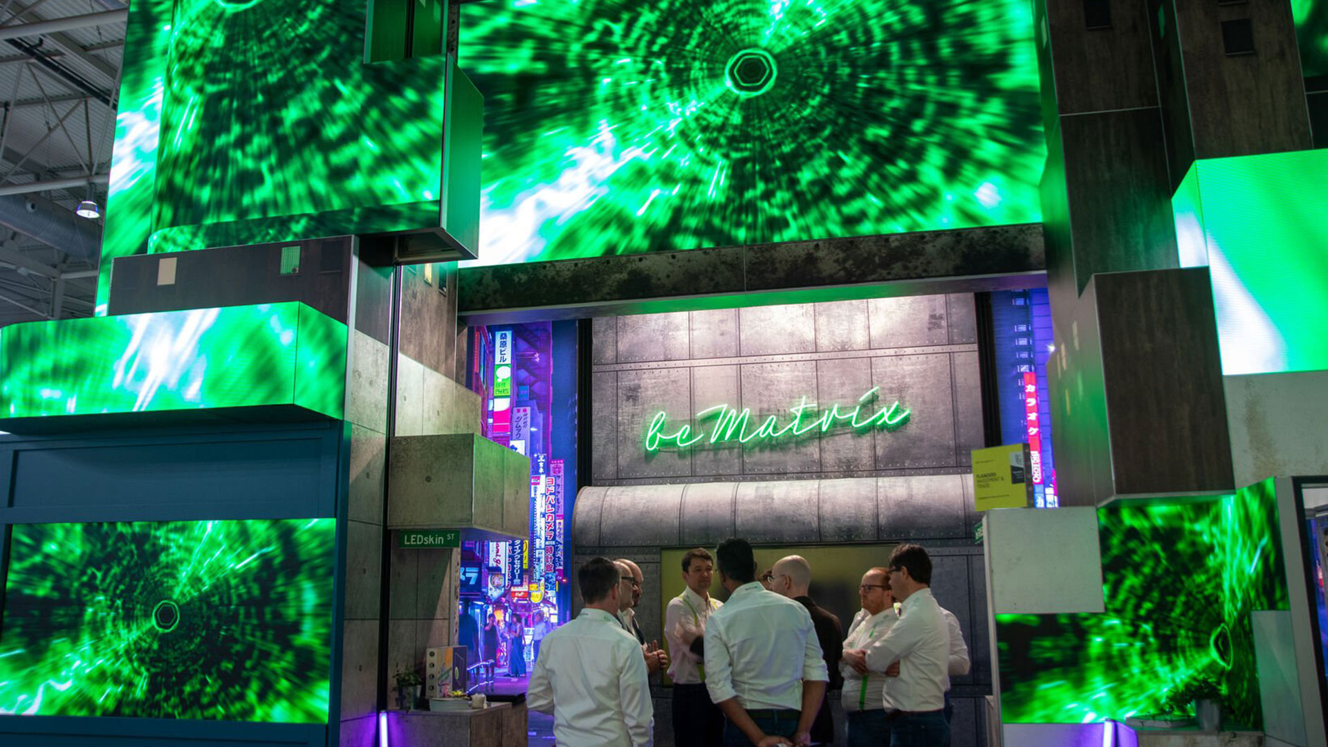 A group of people stand and talk under large green digital screens with swirling matrix-like patterns. The background wall has neon text that reads “be Matrix.” The scene appears to be at a modern trade show or exhibition.