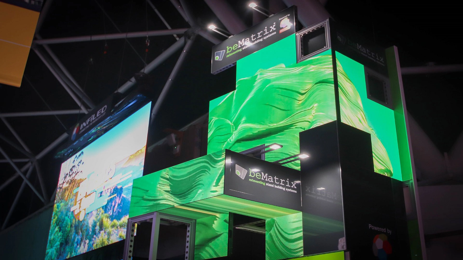 A multi-level exhibition booth features bright green, abstract digital graphics on large LED screens. The booth displays deMatrix logos and is set inside a convention center with other illuminated displays nearby.
