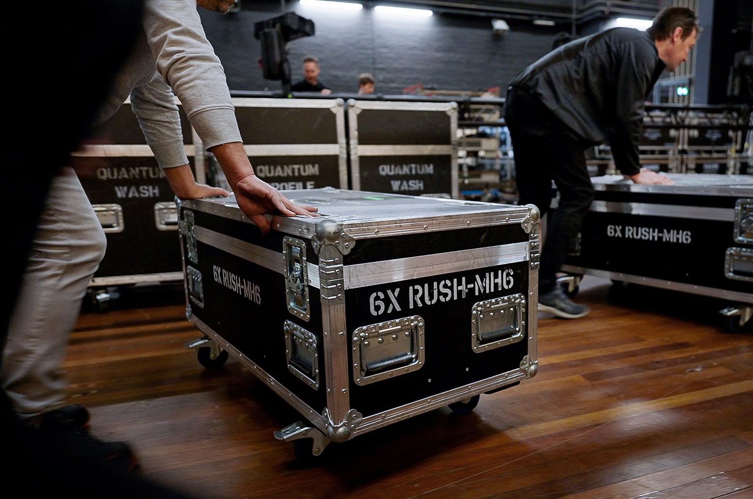People push large black equipment cases labeled 6X RUSH-MH6 across a wooden floor in a backstage or event setup area, with other cases and blurred figures visible in the background.