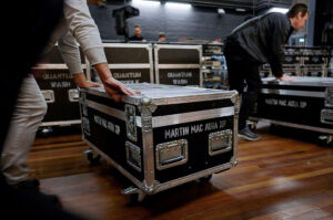 People push large black equipment cases labeled MARTIN MAC AURA XIP and QUANTUM WASH on a wooden floor in a backstage or event setup environment.