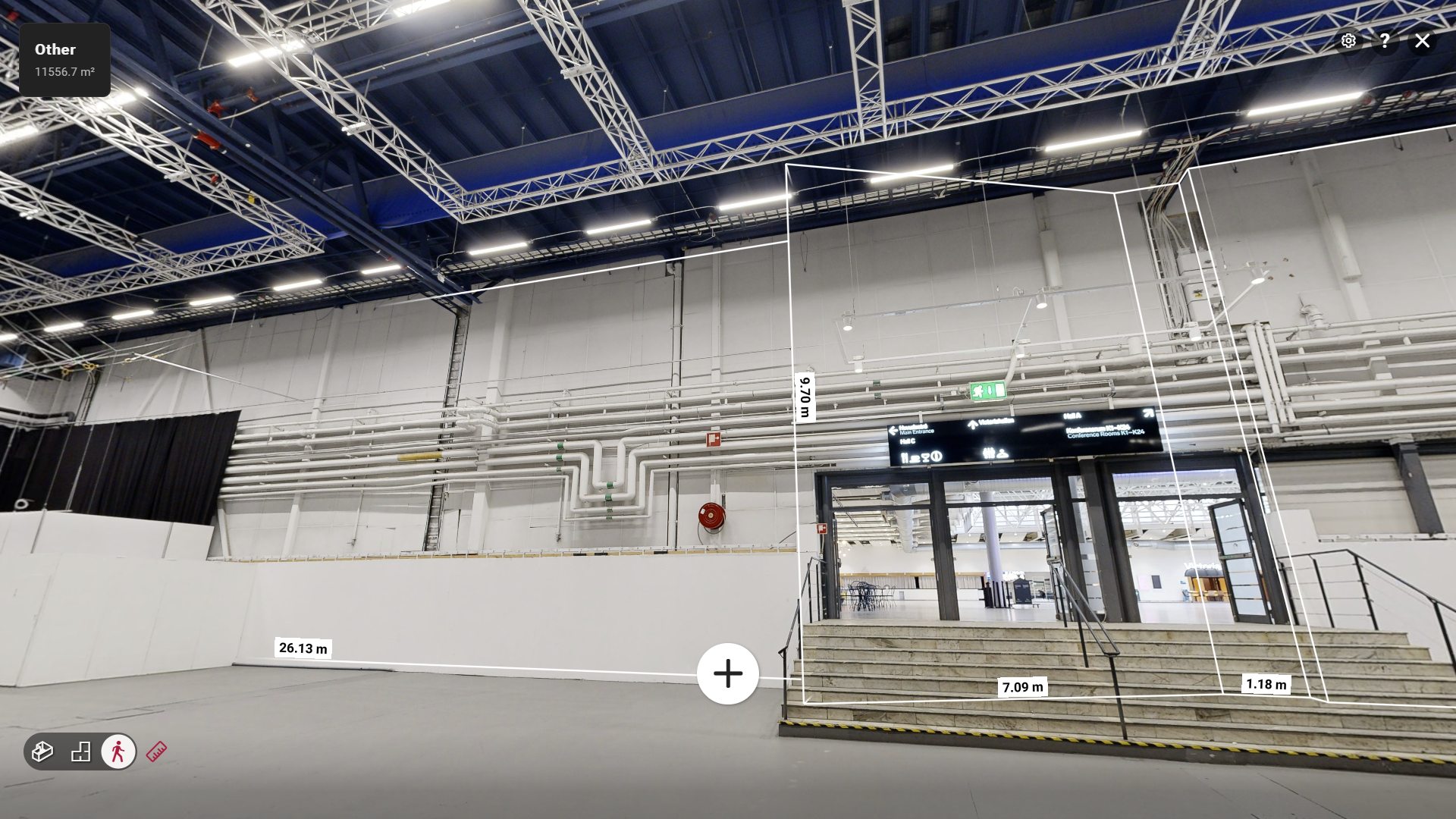 A spacious indoor hall with a high ceiling, visible metal pipes, and overhead lighting. There are stairs leading up to glass doors, emergency exit signs, a fire hose, and measurement lines marking dimensions on the walls and floor.