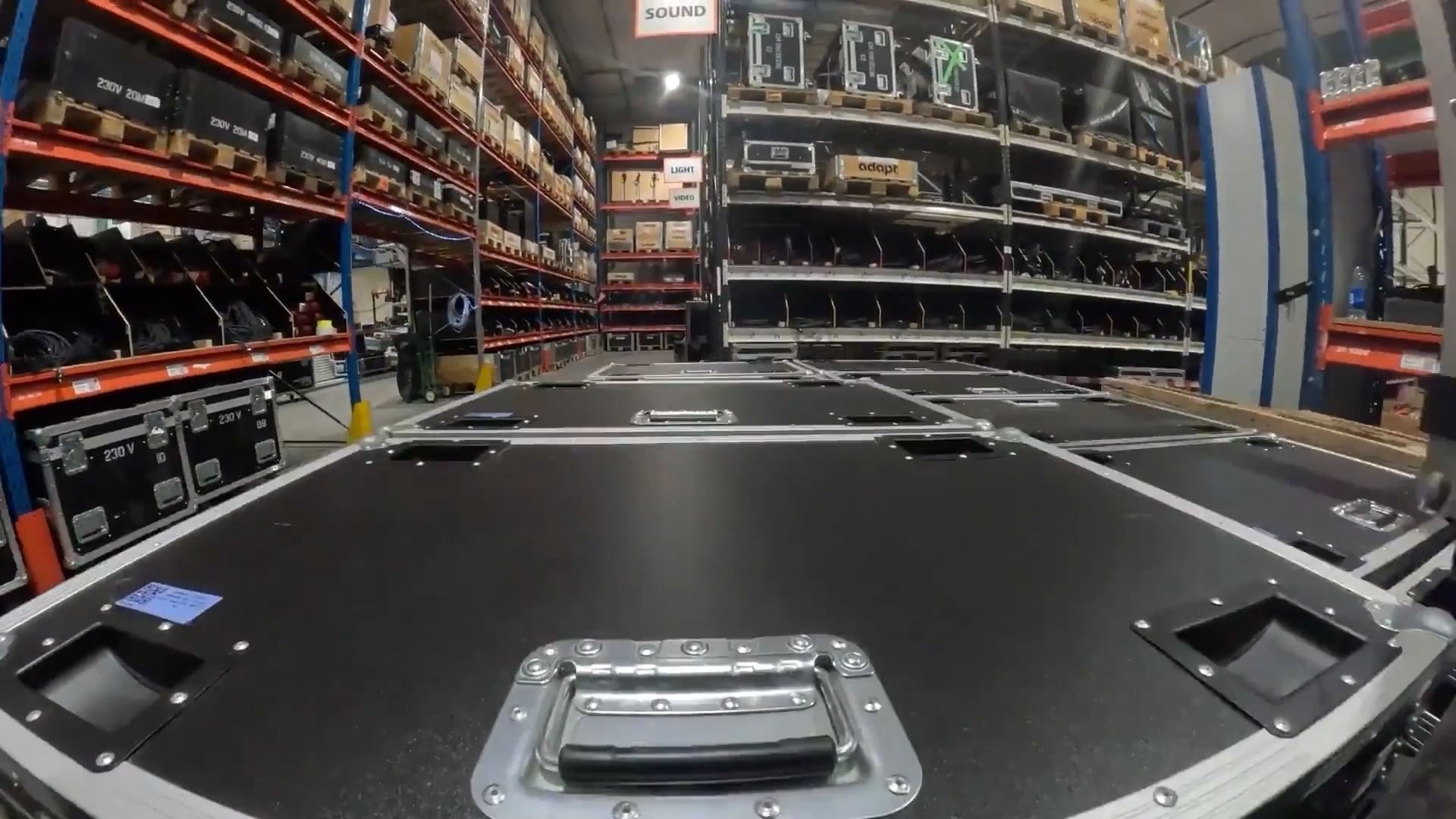 Large, black equipment cases with metal handles are stacked in a warehouse aisle lined with shelves filled with more cases, boxes, and various audio-visual gear. A SOUND sign hangs above the shelves in the background.