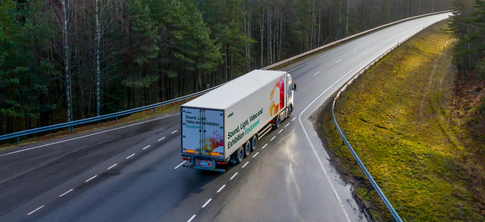 A large white truck drives on an empty, winding highway surrounded by trees and grass on a cloudy day. The trailer features colorful graphics and text about sound, light, and vibration exploration.