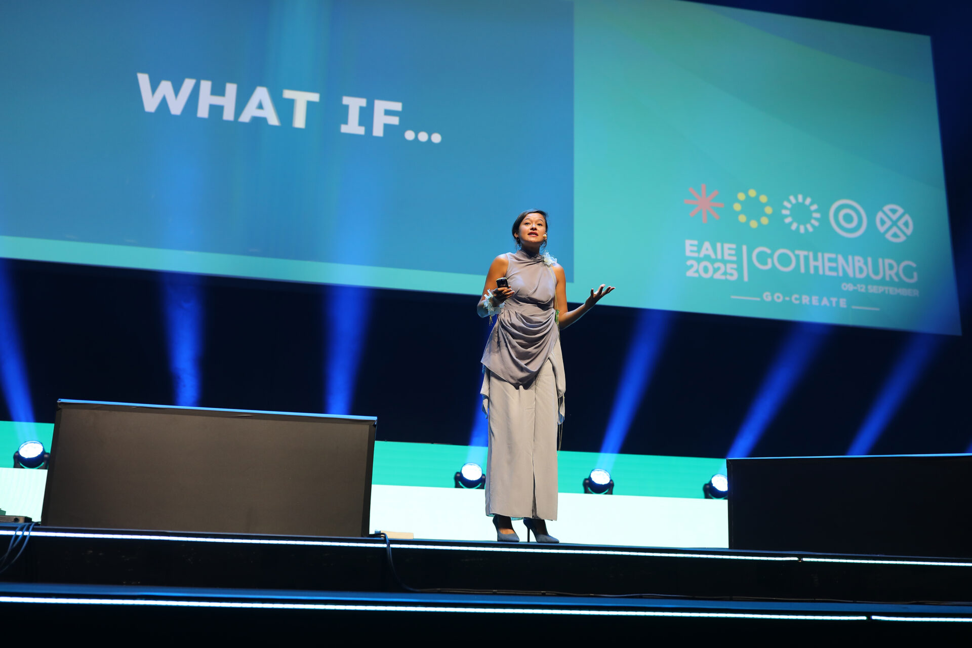 A woman stands on stage speaking in front of a large screen displaying WHAT IF... and the EAIE 2025 Gothenburg Go Create conference logo. Blue and teal lighting highlights the modern setting.
