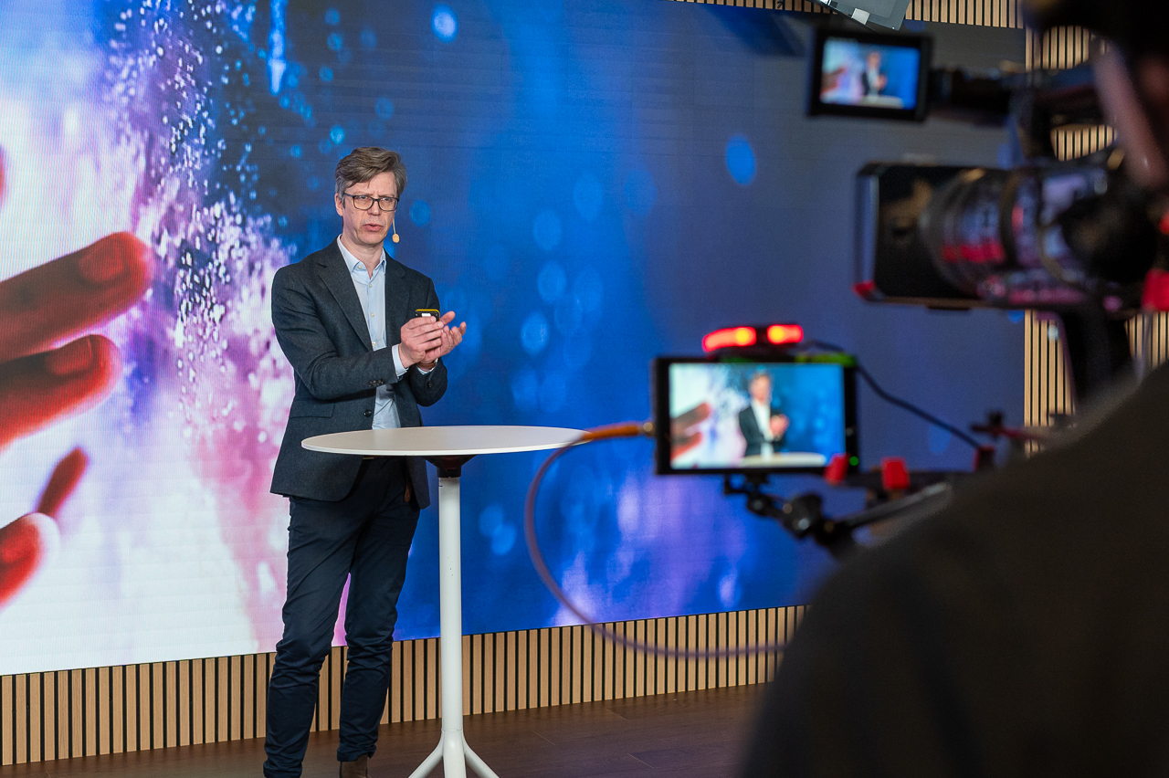 A man in a suit stands at a round table, speaking in front of a large digital screen with abstract graphics, while being filmed by a camera crew.