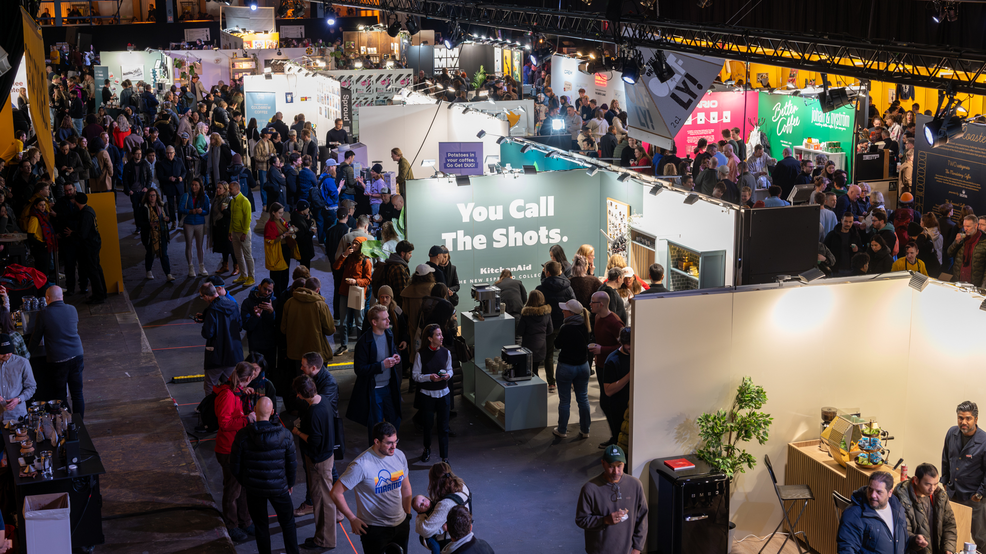 A large indoor event space is filled with people walking among brightly lit booths and displays, including one with a prominent sign that reads You Call The Shots. The atmosphere is lively and bustling.