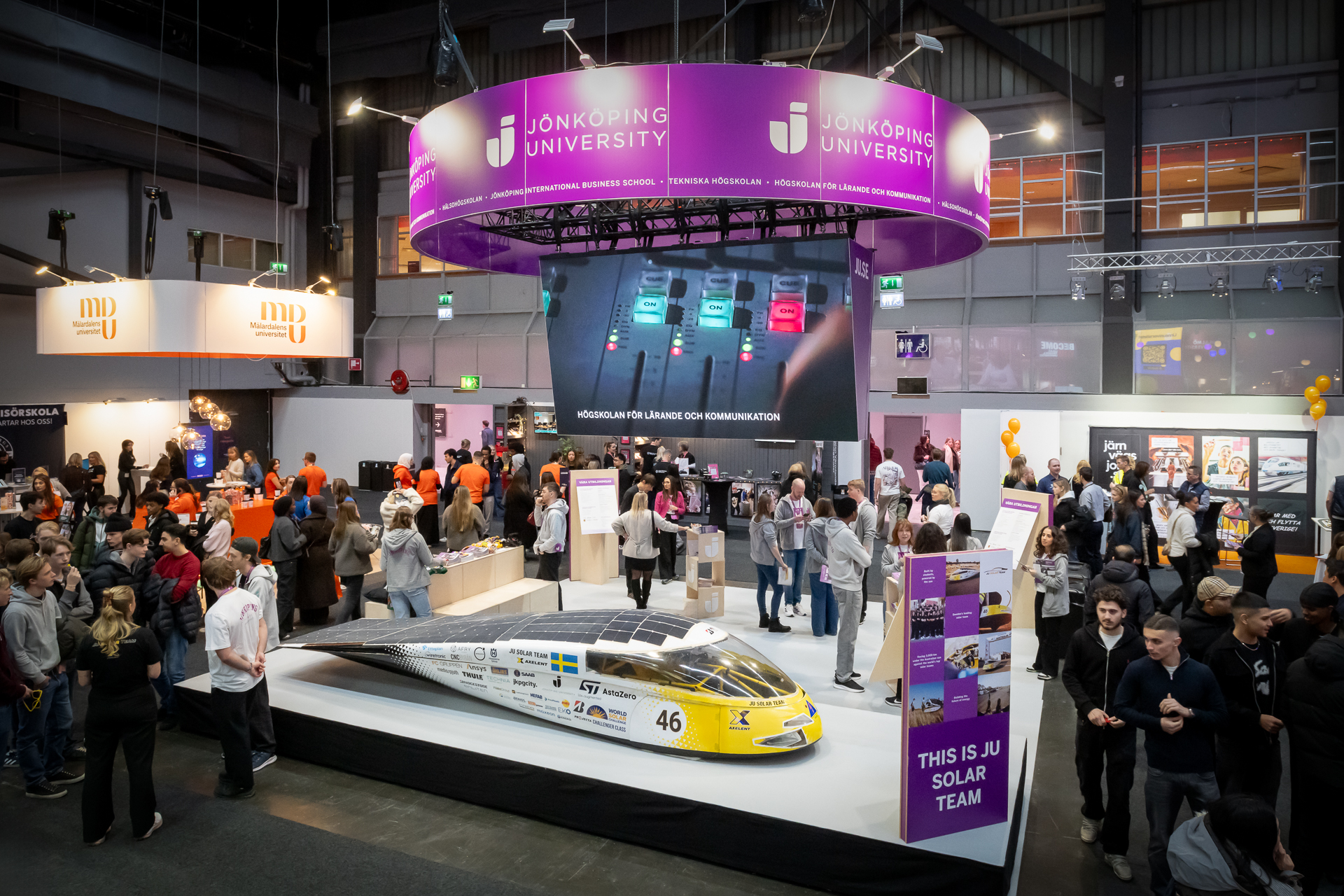 A crowded exhibition hall features a large Jönköping University booth with a solar car on display, informational signs, and a screen above showing digital graphics. Visitors gather around and interact with various exhibits.