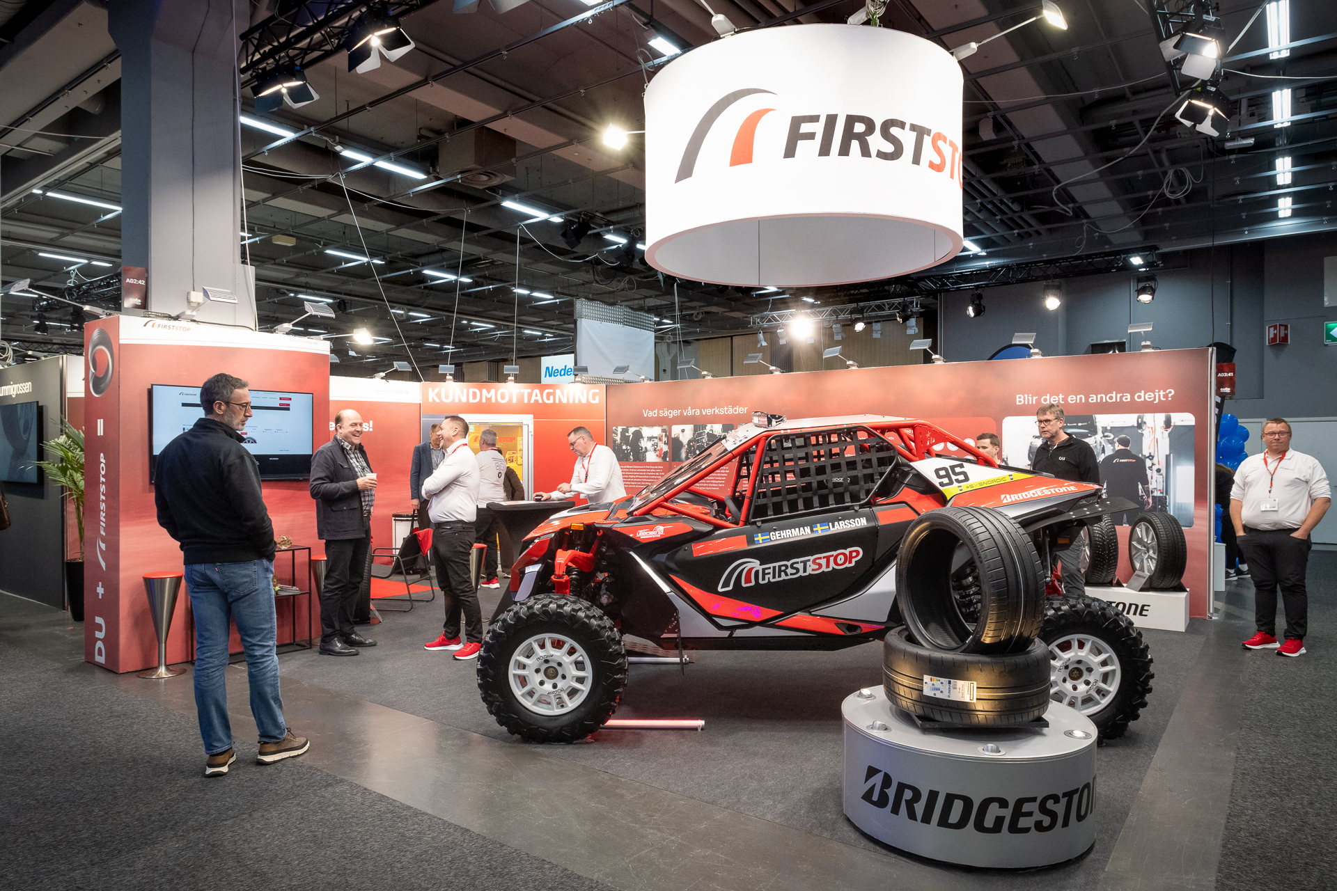 A trade show booth features a red off-road race car with “First Stop” branding, surrounded by tires and display panels. Several people stand and converse near the car under bright lighting.
