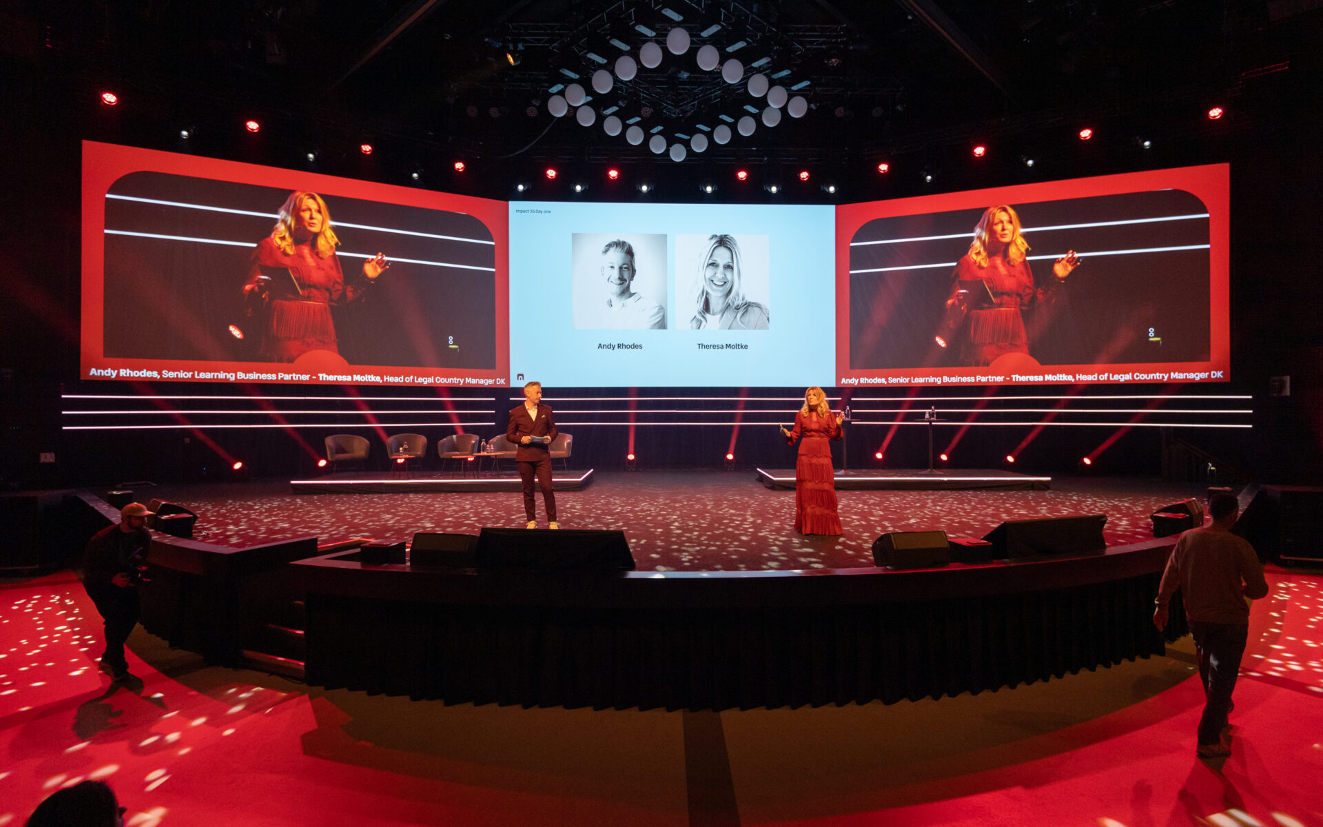 A woman and a man stand on a stage with red lighting, addressing an audience. Behind them, large screens display portraits and names of two people. The stage is spacious, with empty chairs and a few people in the foreground.