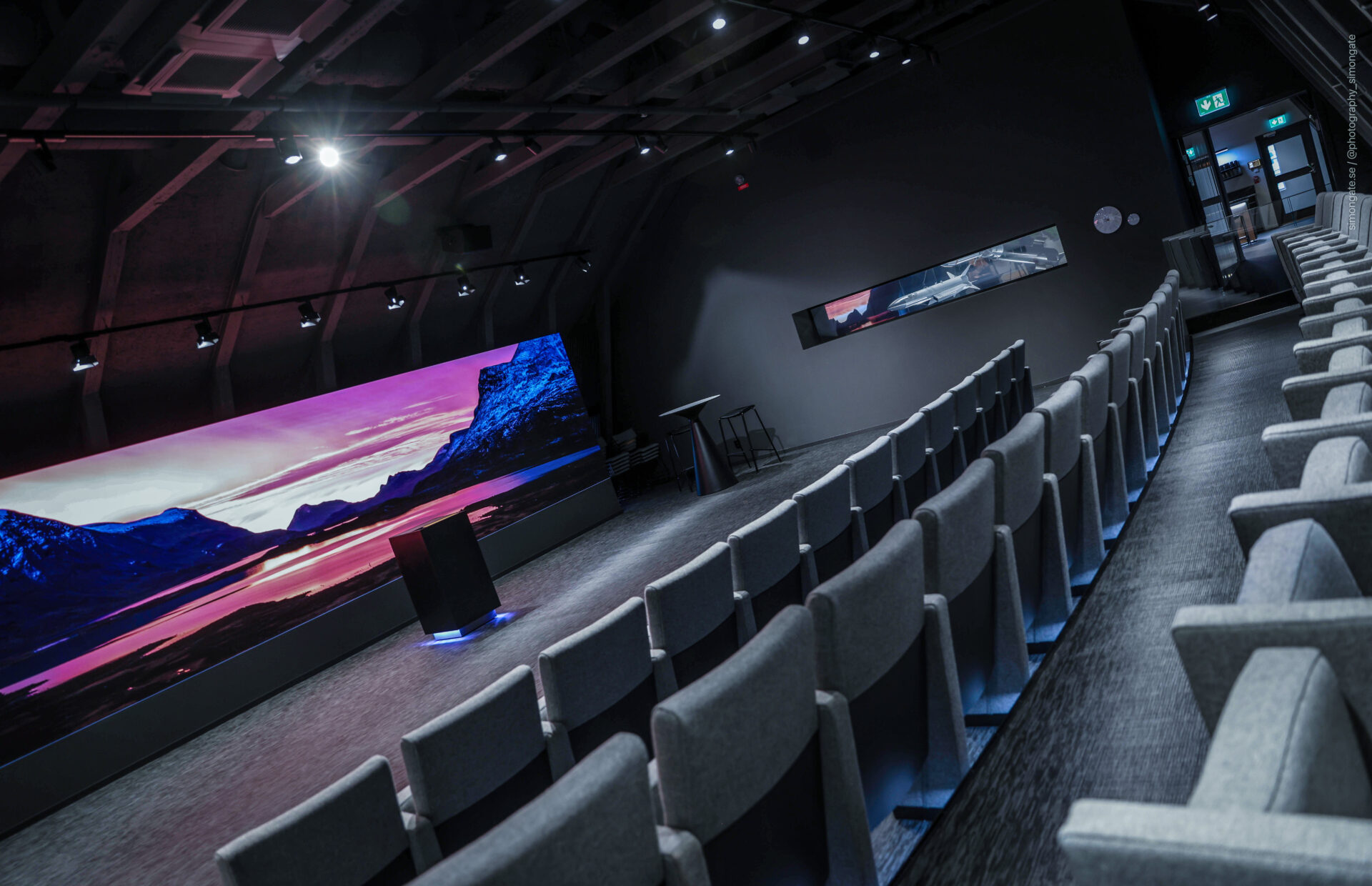 A modern, empty auditorium with rows of gray seats, a large curved screen displaying a scenic mountain landscape, and spotlights illuminating the space. A doorway and a long mirror are visible on the dark walls.