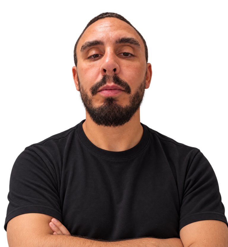 A man with short hair and a beard wears a black t-shirt and stands with arms crossed, looking confidently at the camera against a plain white background.