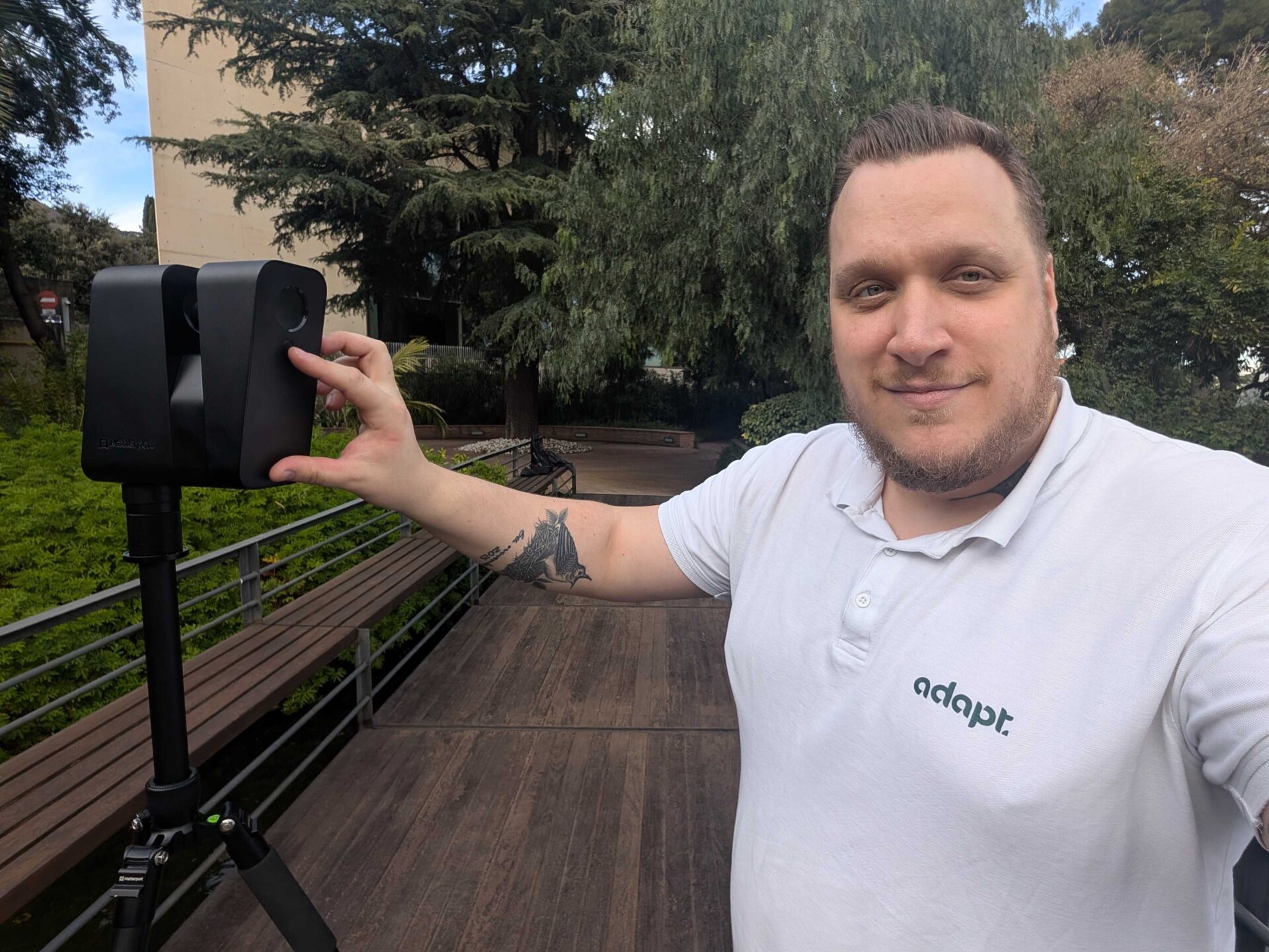 A person in a white adapt. polo shirt stands outdoors on a wooden walkway, smiling and holding a black device on a tripod with greenery and trees in the background.
