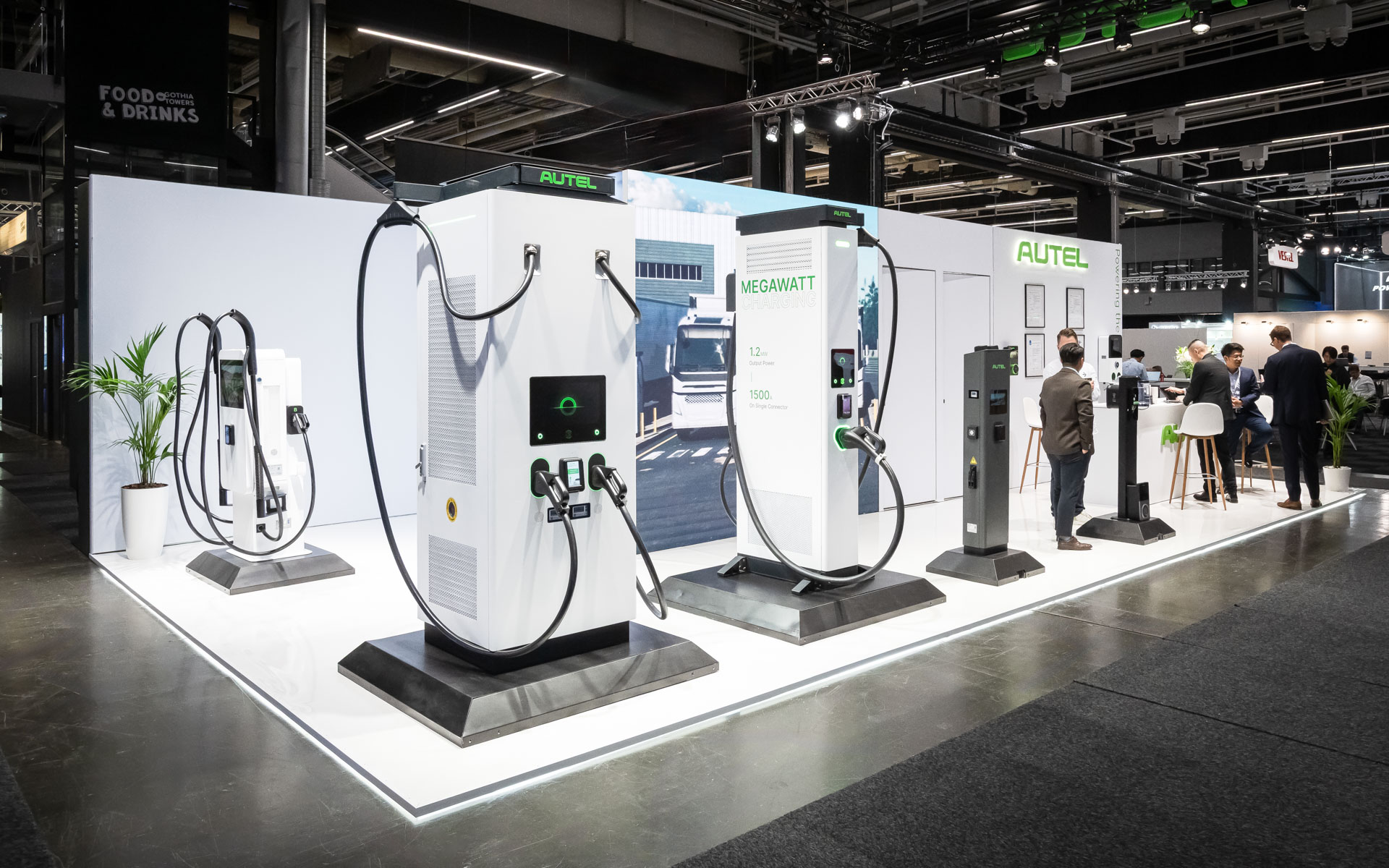 A trade show booth displays several types of electric vehicle charging stations, with people standing and conversing nearby. The booth is modern, well-lit, and features branding by AUTEL and MEGAWATT.