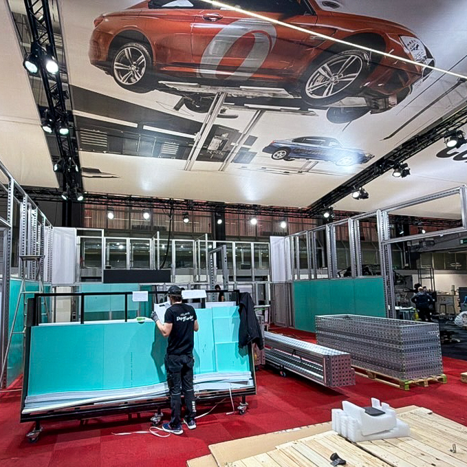 A person works on assembling a turquoise display structure in an exhibition hall with red carpet. Above, a ceiling banner shows a large image of a red sports car. Metal frames and packing materials are scattered around.