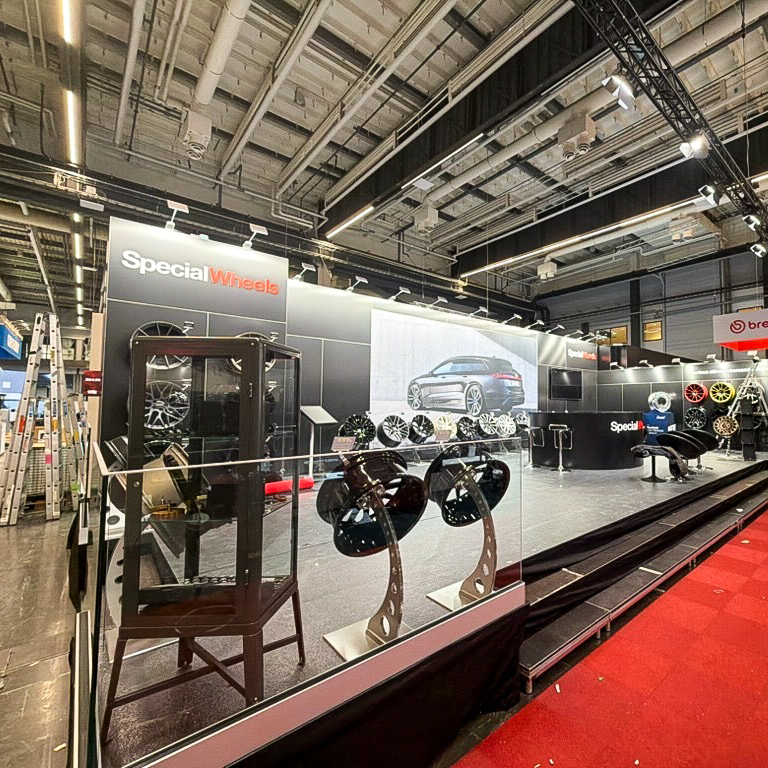 A trade show booth for Special Wheels displays various car rims and wheels, with a large car image on the wall and glass cases showcasing products under bright, industrial lighting.