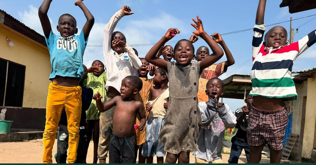 A group of children outdoors joyfully jump and raise their arms, smiling and laughing together under a bright blue sky near yellow buildings.