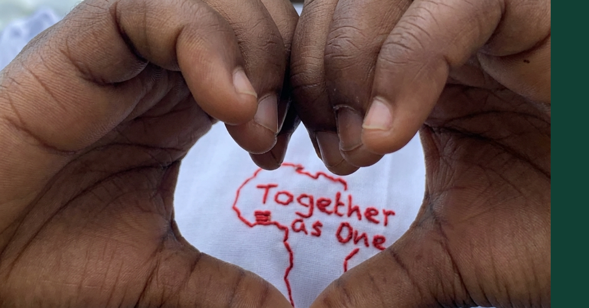 Two hands form a heart shape in front of a shirt embroidered with an outline of Africa and the words “Together as One” in red thread.