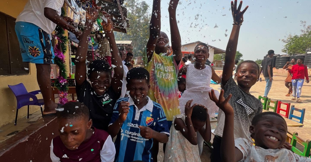 A group of joyful children outdoors laugh and throw confetti into the air at a celebration, with colorful chairs and decorations in the background on a sunny day.