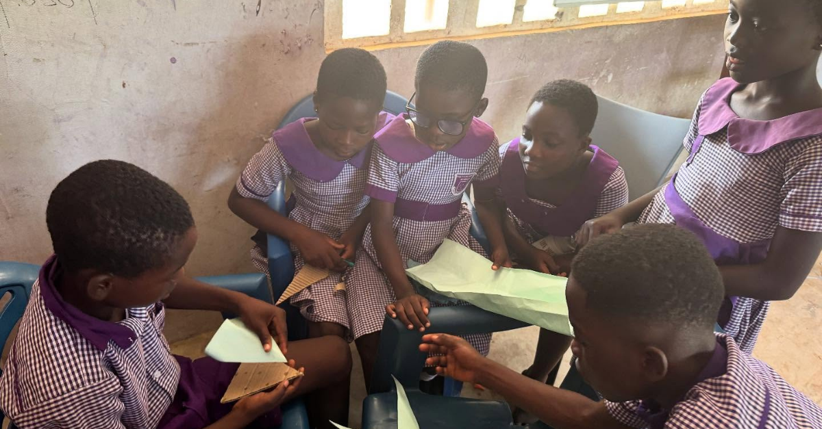 A group of school children in purple checkered uniforms sit closely together, folding paper and engaging in a classroom activity. They appear focused and are seated on blue plastic chairs near a sunlit window.