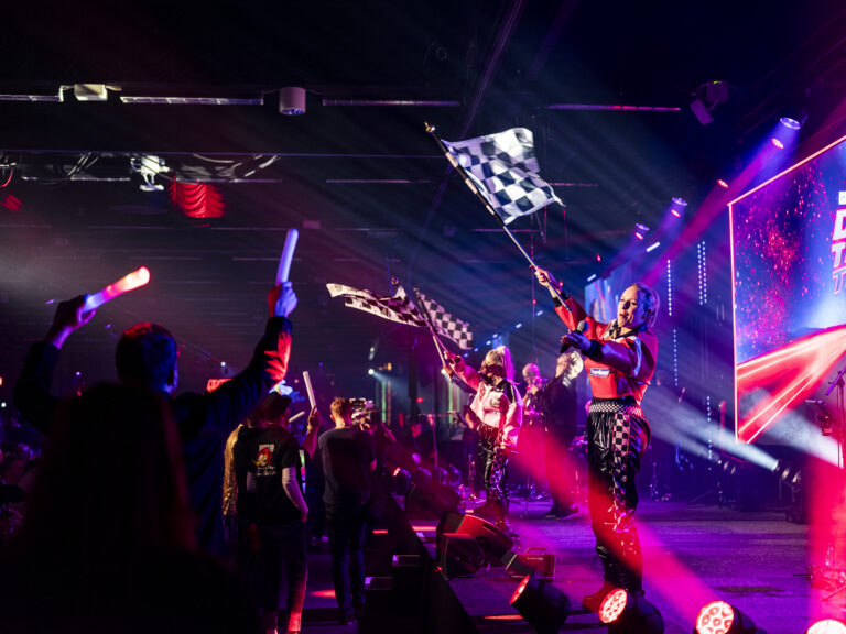 Performers in racing outfits wave checkered flags on a brightly lit stage, while an enthusiastic crowd waves glowing sticks. Colorful lights and a large screen create an energetic concert atmosphere.