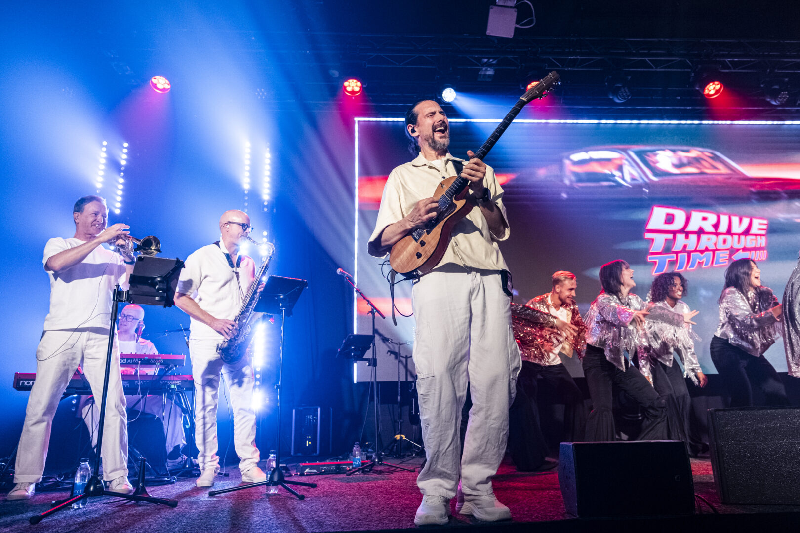 A band dressed in white performs energetically on stage, featuring a guitarist in the foreground and brass musicians. Behind them, backup singers dance in silver outfits, with a large “Drive Through Time” sign on a screen in the background.