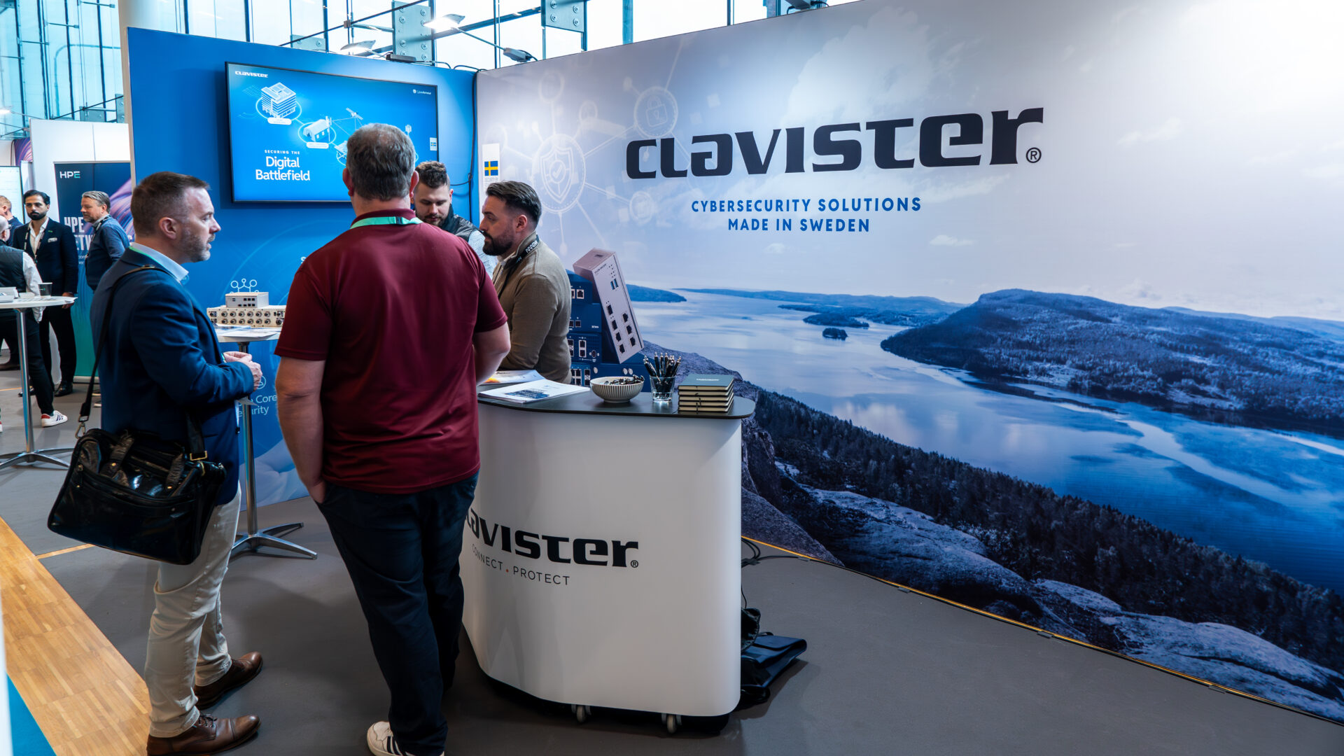 Four men converse at a Clavister booth at a tech expo. The booth displays a large scenic backdrop of a Swedish lake and forest, and signage reads CYBERSECURITY SOLUTIONS MADE IN SWEDEN. Tech equipment and brochures are on the counter.