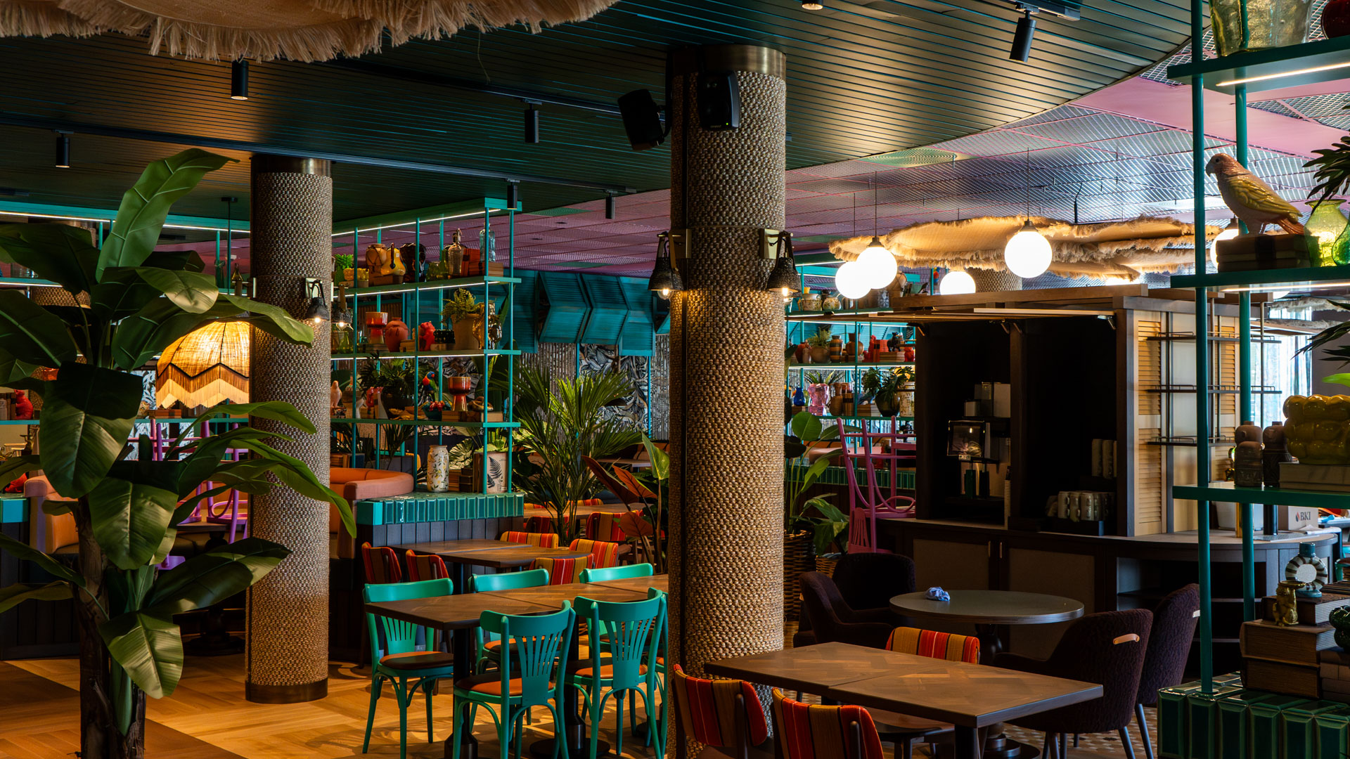 A vibrant, modern restaurant interior with colorful chairs, patterned pillars, lush indoor plants, and shelves displaying eclectic decor under warm, ambient lighting.