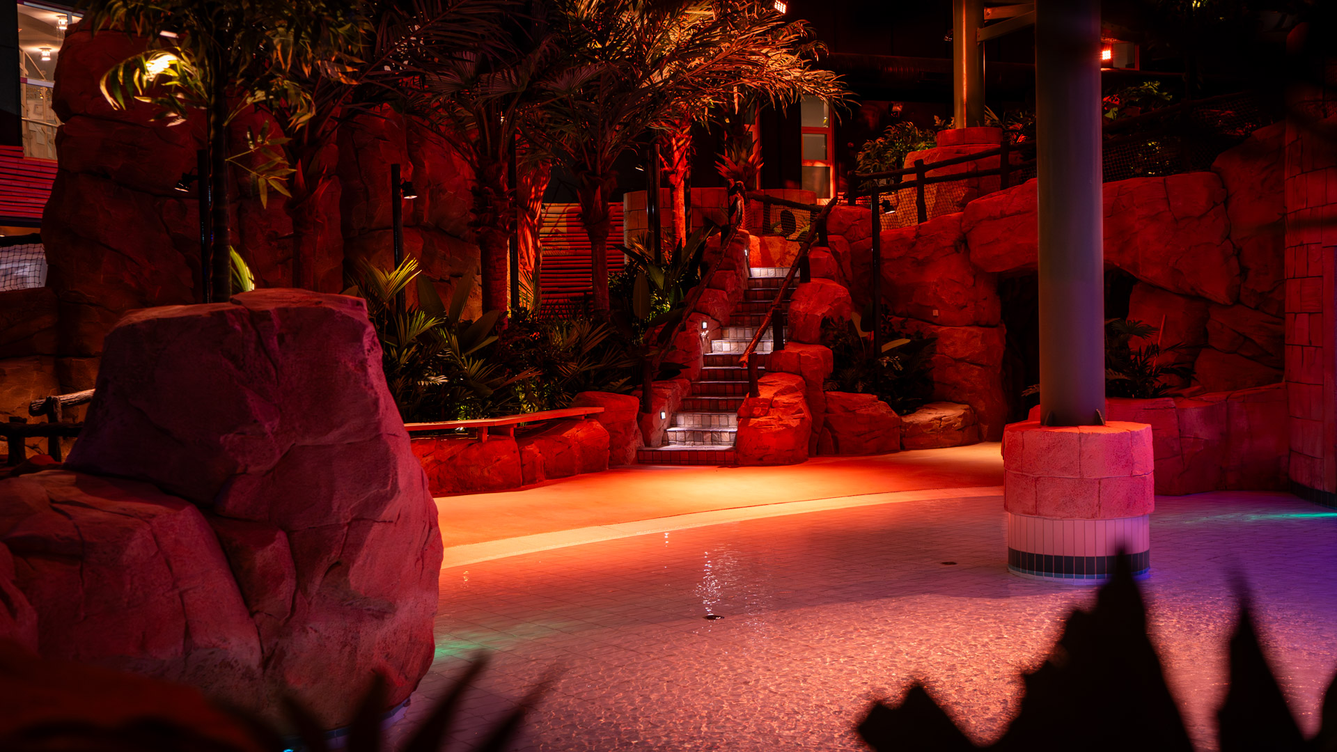 An indoor pool area with artificial rocks, tropical plants, and illuminated stairs leading upwards. Warm, red and orange lighting creates a tropical, exotic atmosphere. The pool is shallow and calm.