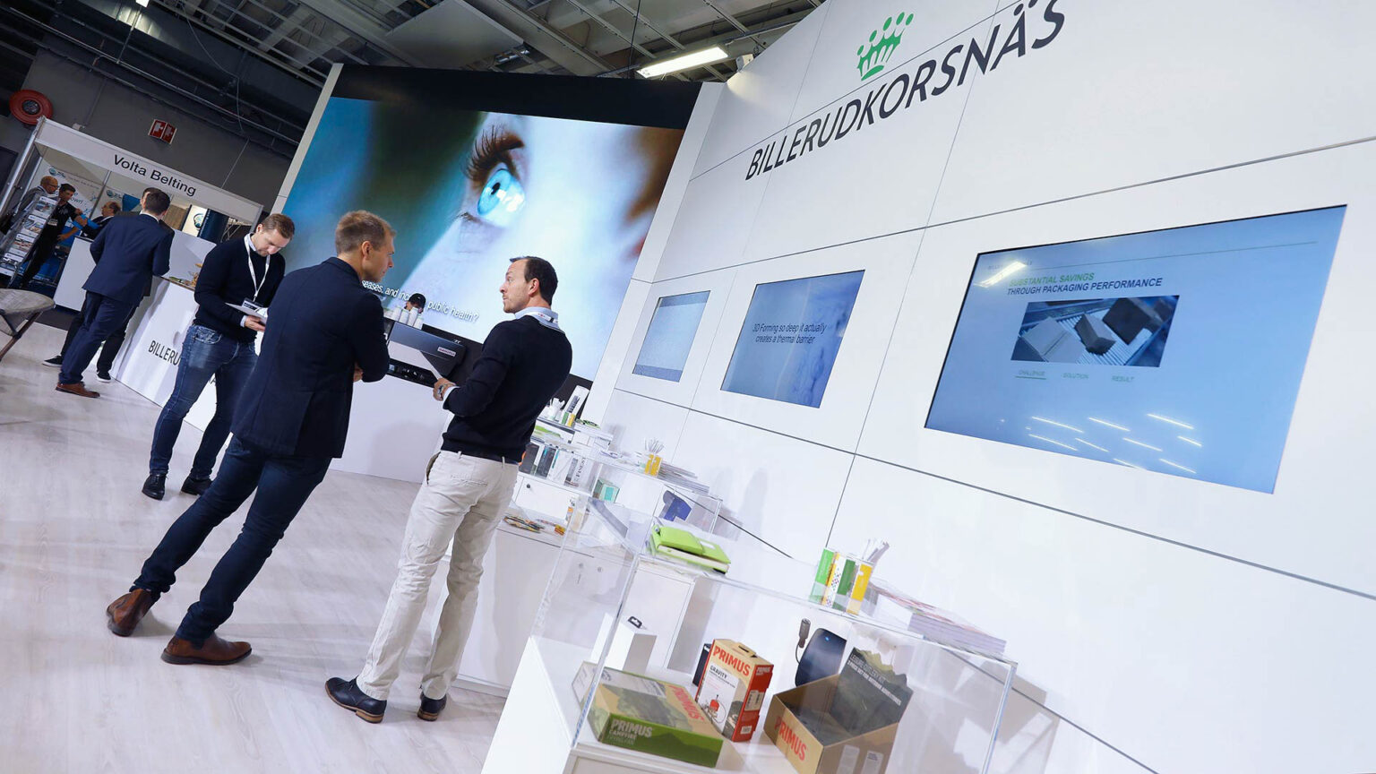 People interact at a modern exhibition booth for BillerudKorsnäs, featuring large digital screens, a display counter with product samples, and a futuristic eye graphic in the background.