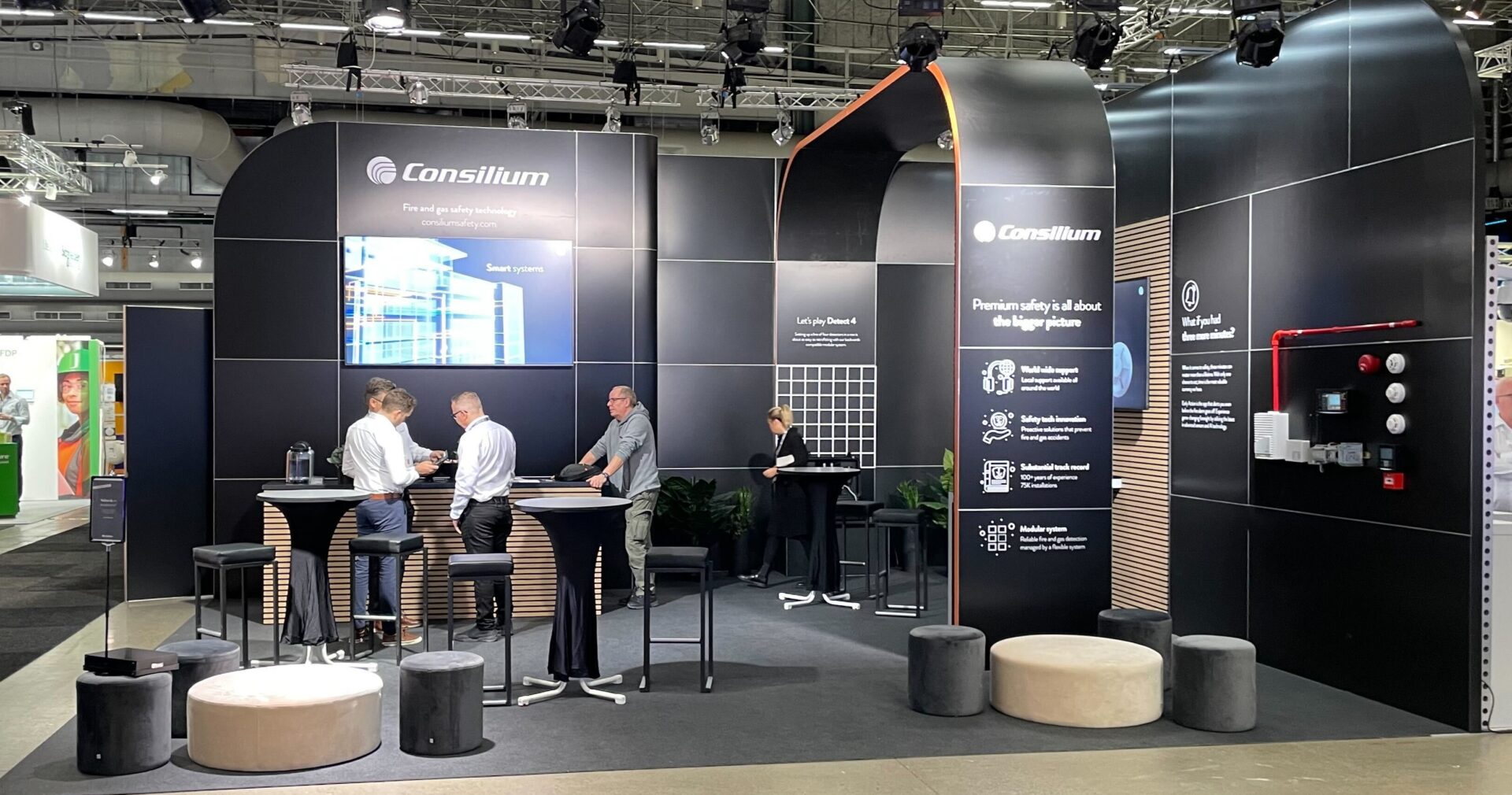 A modern trade show booth for Consilium features four people talking and standing around high tables, with displays, product panels, stools, and black walls showcasing company information and graphics.